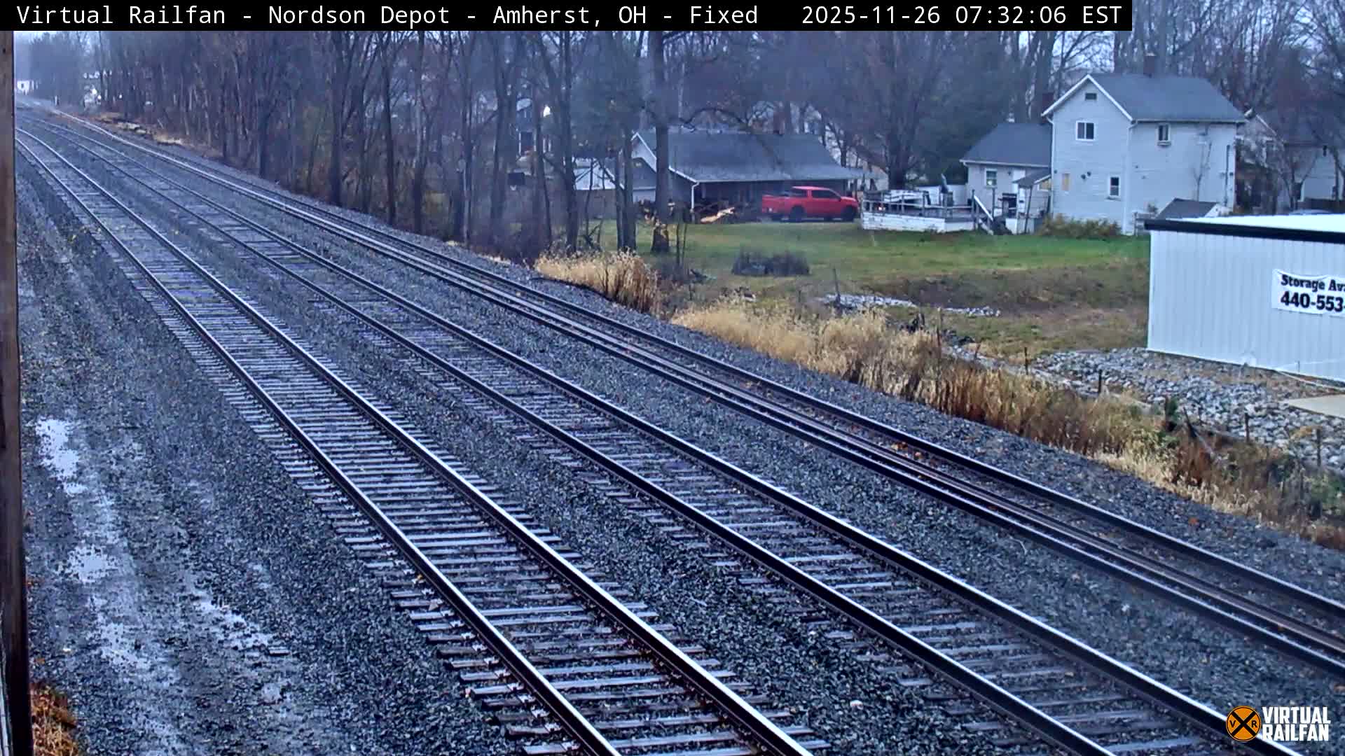 Multiple railroad tracks lead into the distance alongside an overgrown area, with dimly lit buildings and a storage container visible in the background under a clear night sky.