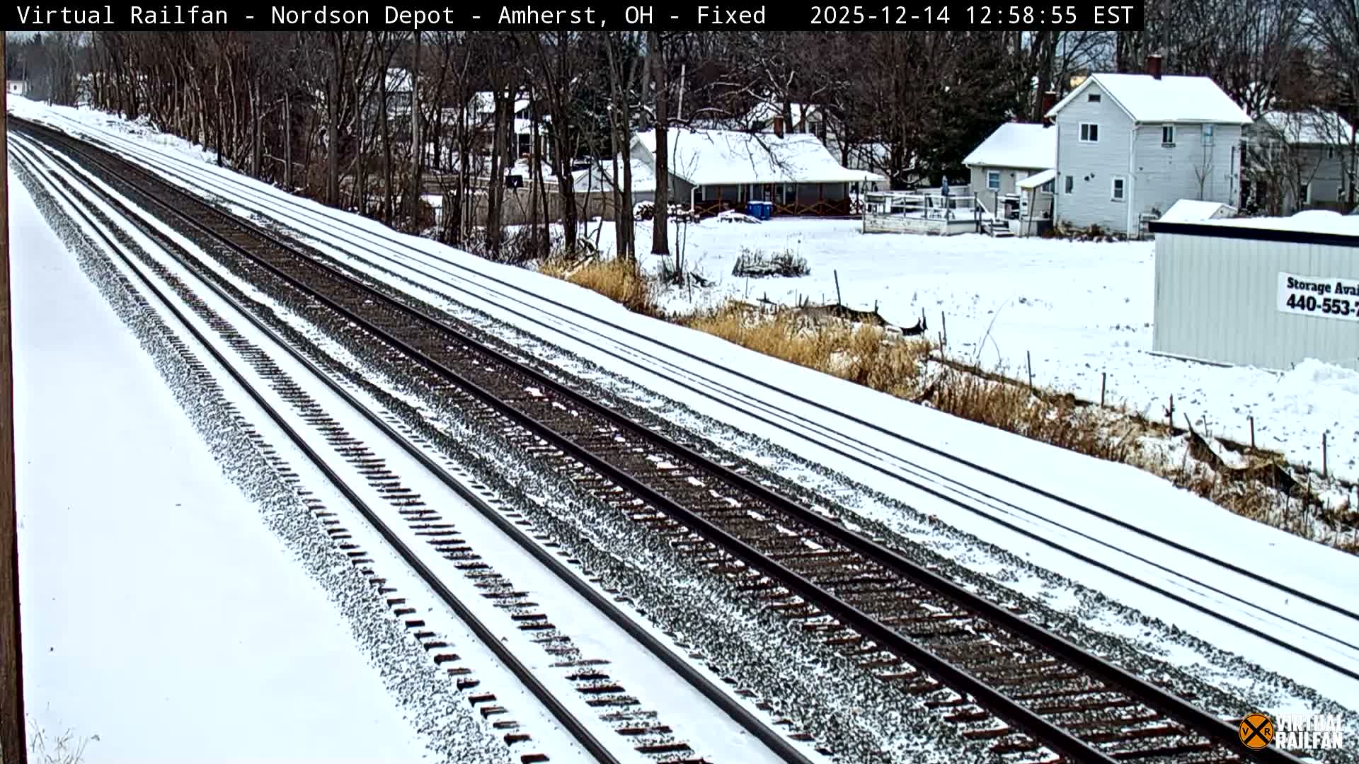 Multiple railroad tracks lead into the distance alongside an overgrown area, with dimly lit buildings and a storage container visible in the background under a clear night sky.