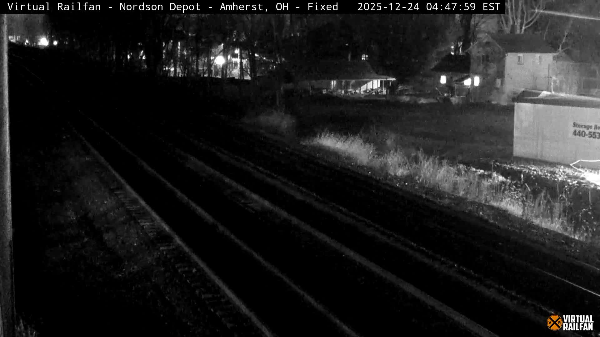 Multiple railroad tracks lead into the distance alongside an overgrown area, with dimly lit buildings and a storage container visible in the background under a clear night sky.