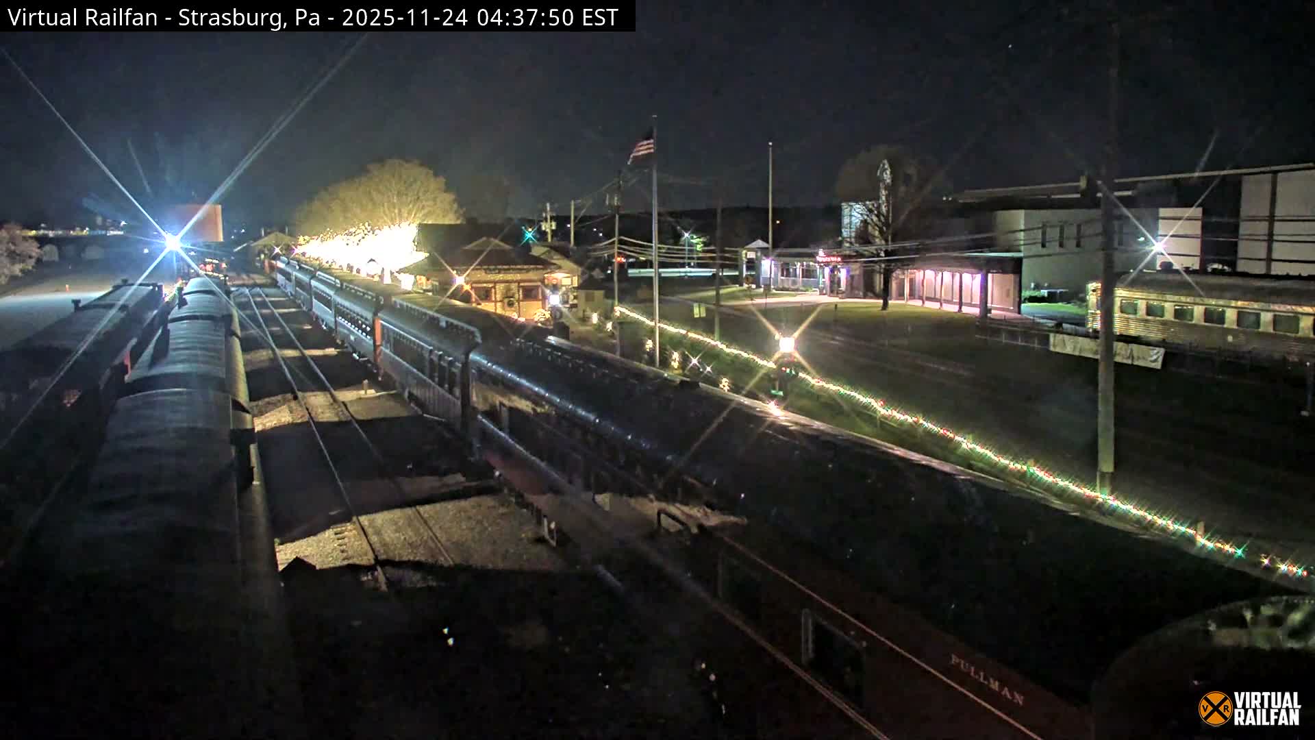 The image presents a night scene at a train station under a clear sky, showcasing multiple trains on parallel tracks, brightly lit station buildings, and a long string of decorative lights lining the trackside.