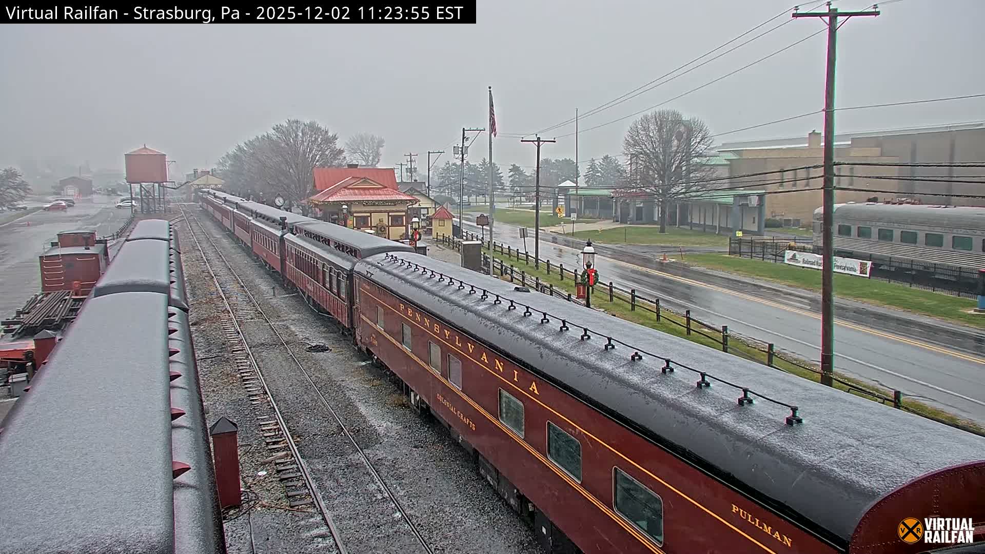 A long maroon passenger train with visible branding like "PENNSYLVANIA" and "PULLMAN" sits on wet tracks outside a station, amidst a light covering of wet snow or sleet under an overcast sky.