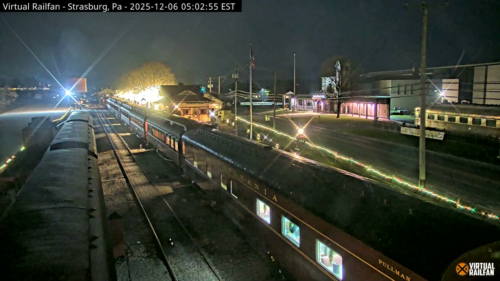 A passenger train with illuminated windows and festive string lights is parked at a brightly lit station under a clear night sky.