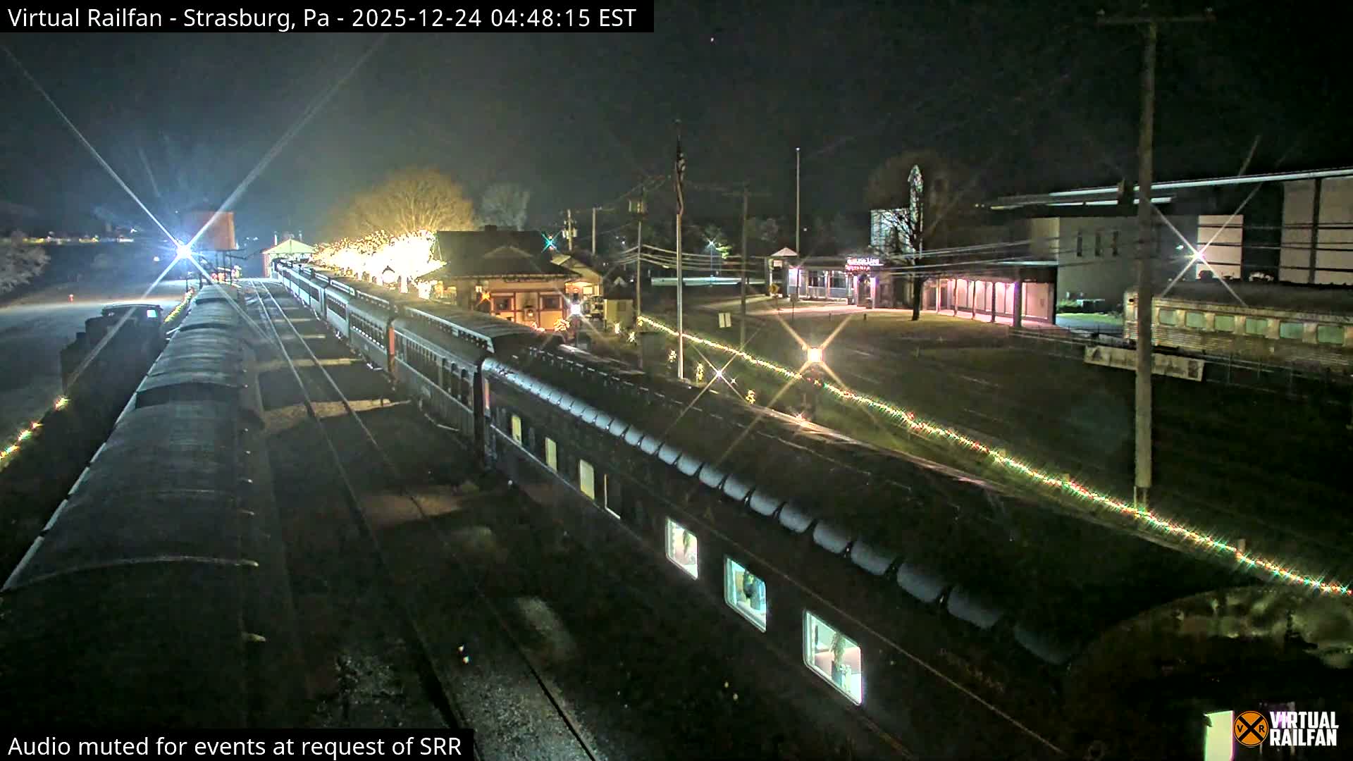 A passenger train with illuminated windows and festive string lights is parked at a brightly lit station under a clear night sky.