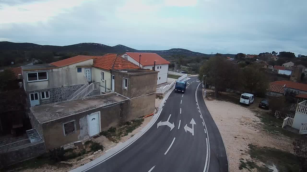 An elevated view captures a village scene with a blue truck driving on a winding road marked with turning arrows, surrounded by houses with red tiled roofs and distant hills under an overcast sky.