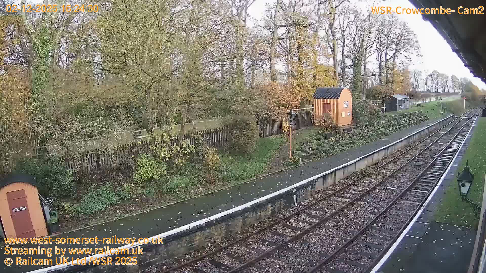 On an overcast and damp day, a rural railway station features two tracks alongside a wet platform, bordered by autumn-toned trees and two small utility buildings.