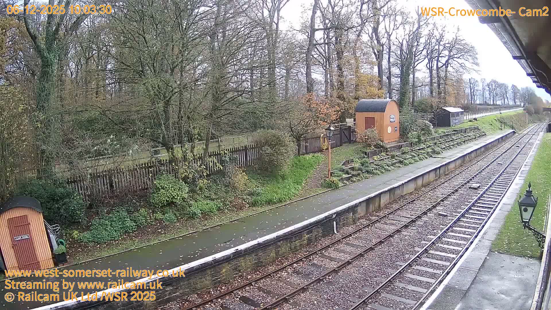 A damp and overcast day reveals a quiet rural railway station with multiple wet tracks, two platforms, small utility buildings, and a backdrop of mostly bare trees and sparse autumn foliage.