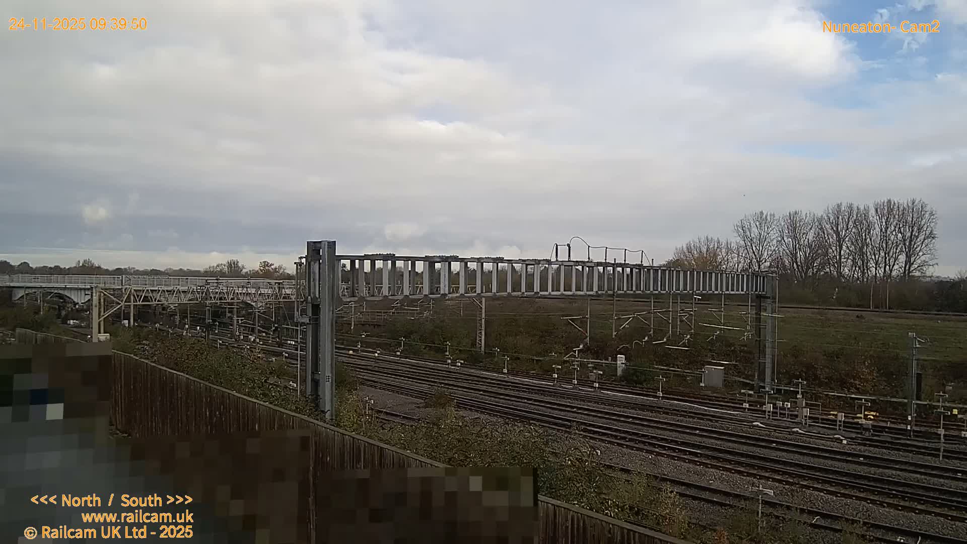 A wide view of a railway junction features numerous tracks, overhead lines, and signal gantries, set against a backdrop of a large bridge and bare trees under a partly cloudy sky.
