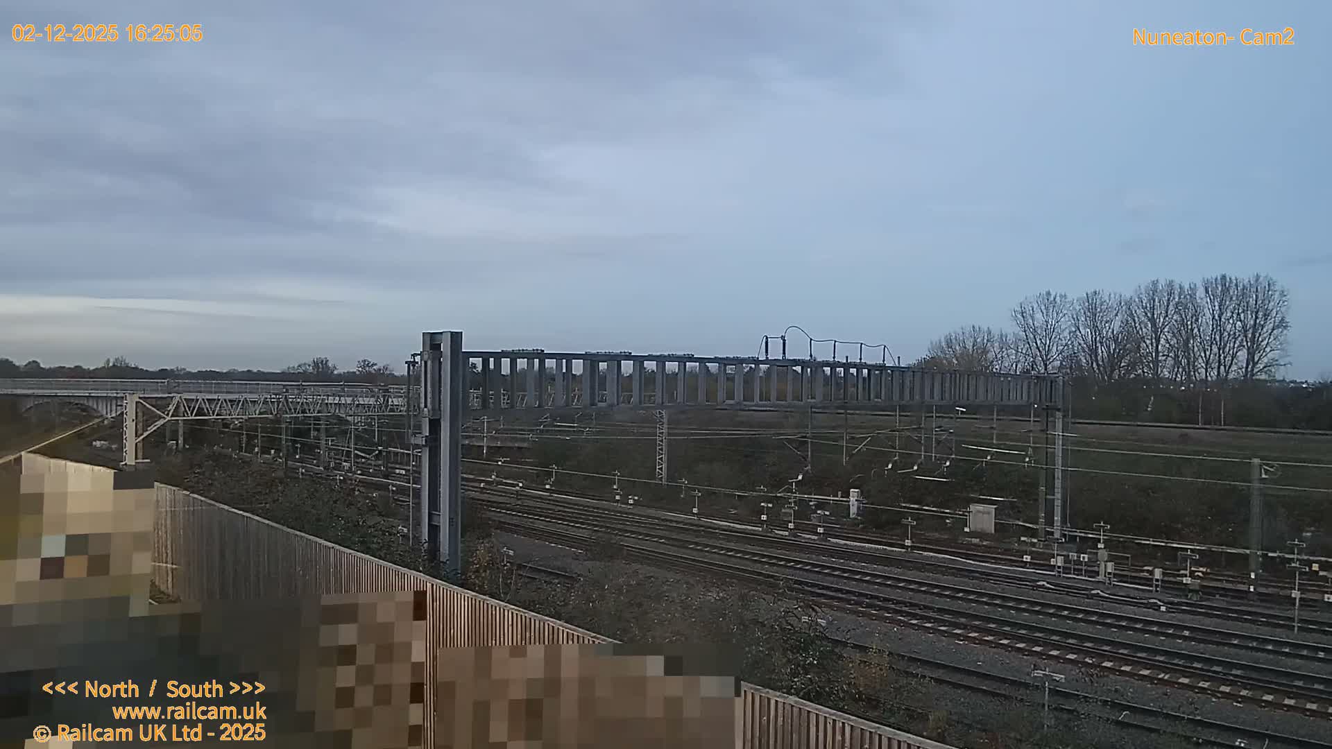 The image depicts a complex railway network with multiple tracks and overhead line equipment, viewed under an overcast grey sky on a dull day, with a long bridge and bare trees visible in the distance.