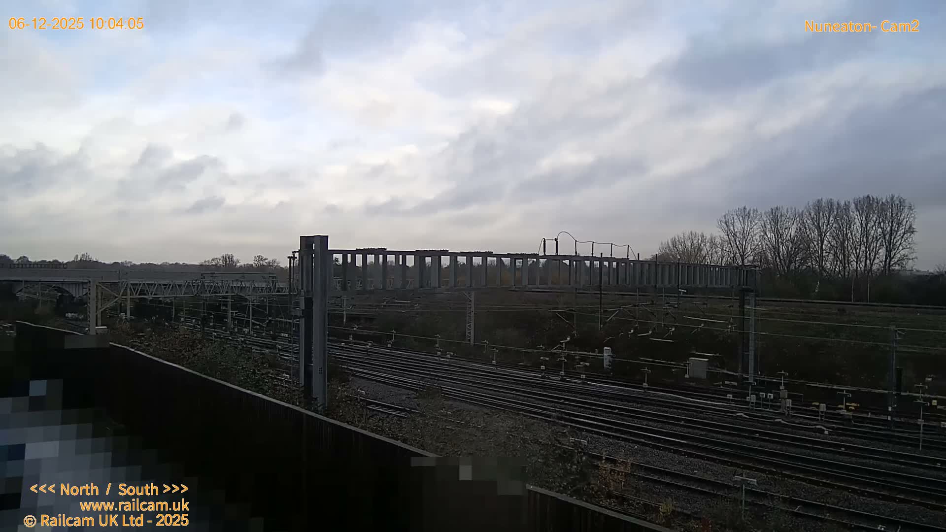 Multiple train tracks with a complex network of overhead gantries stretch into the distance, framed by a treeline on the horizon, under a heavily overcast sky.