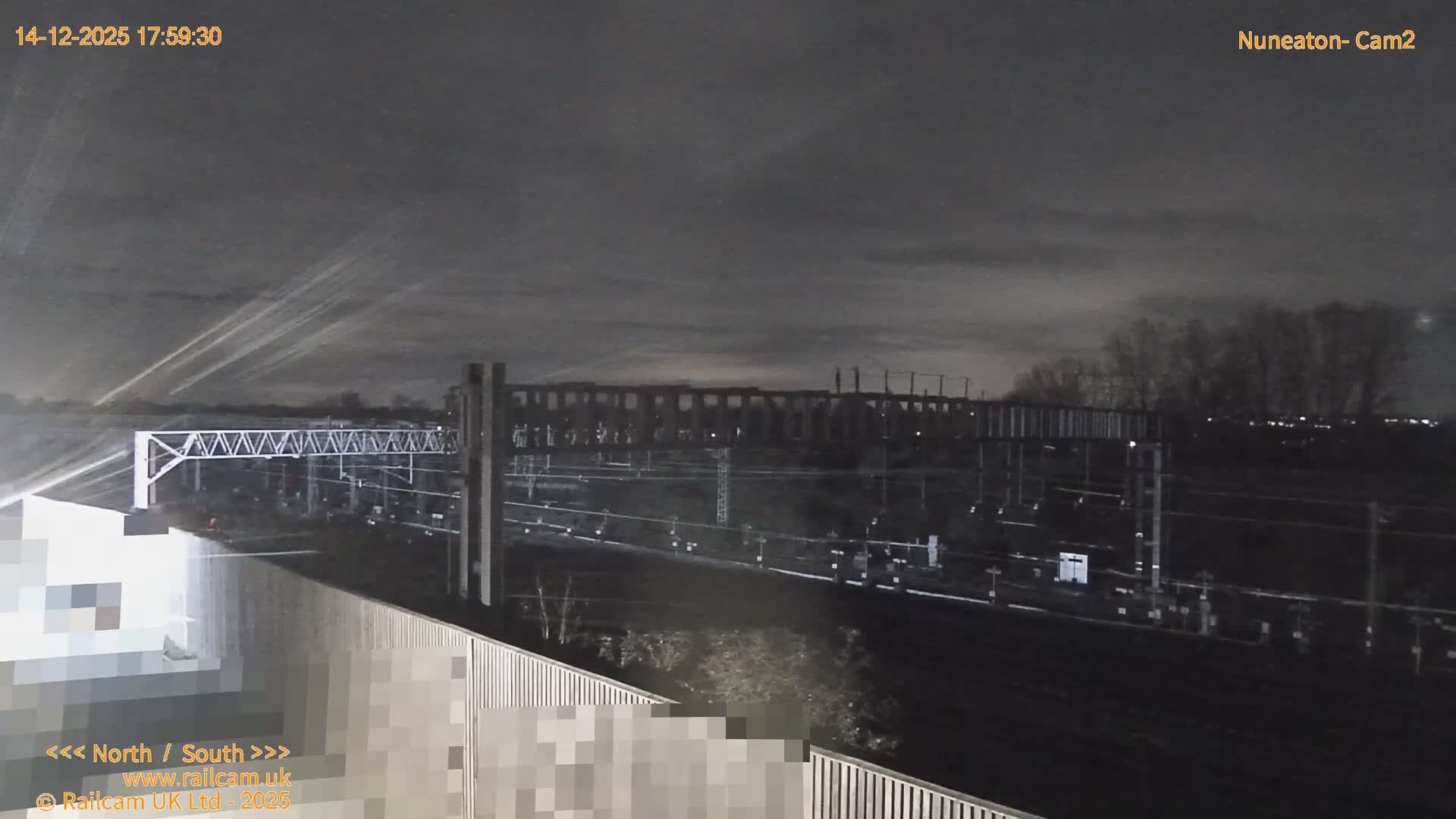 Multiple train tracks with a complex network of overhead gantries stretch into the distance, framed by a treeline on the horizon, under a heavily overcast sky.