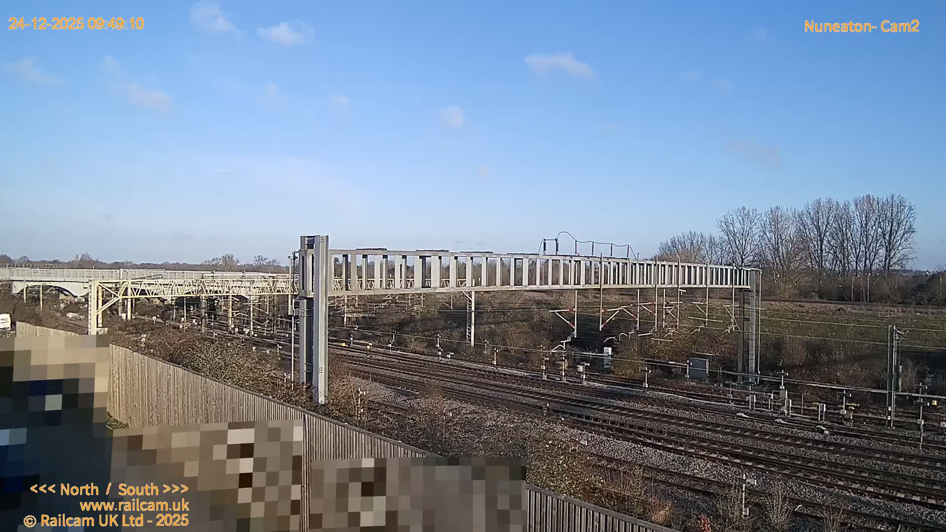 Multiple train tracks with a complex network of overhead gantries stretch into the distance, framed by a treeline on the horizon, under a heavily overcast sky.