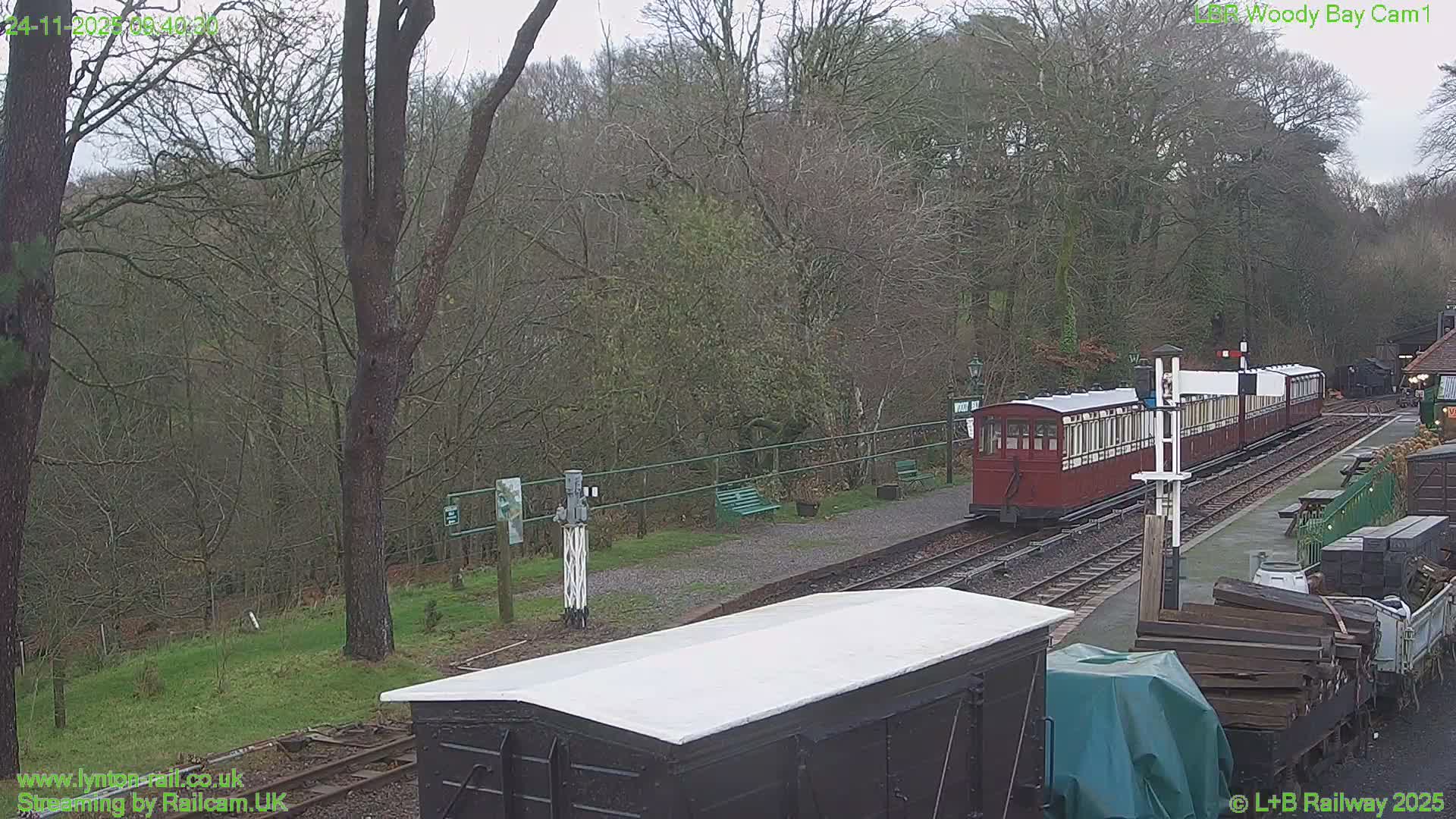 A red and white train with multiple carriages is parked at a rustic outdoor railway station, surrounded by bare trees and railway equipment under an overcast sky.