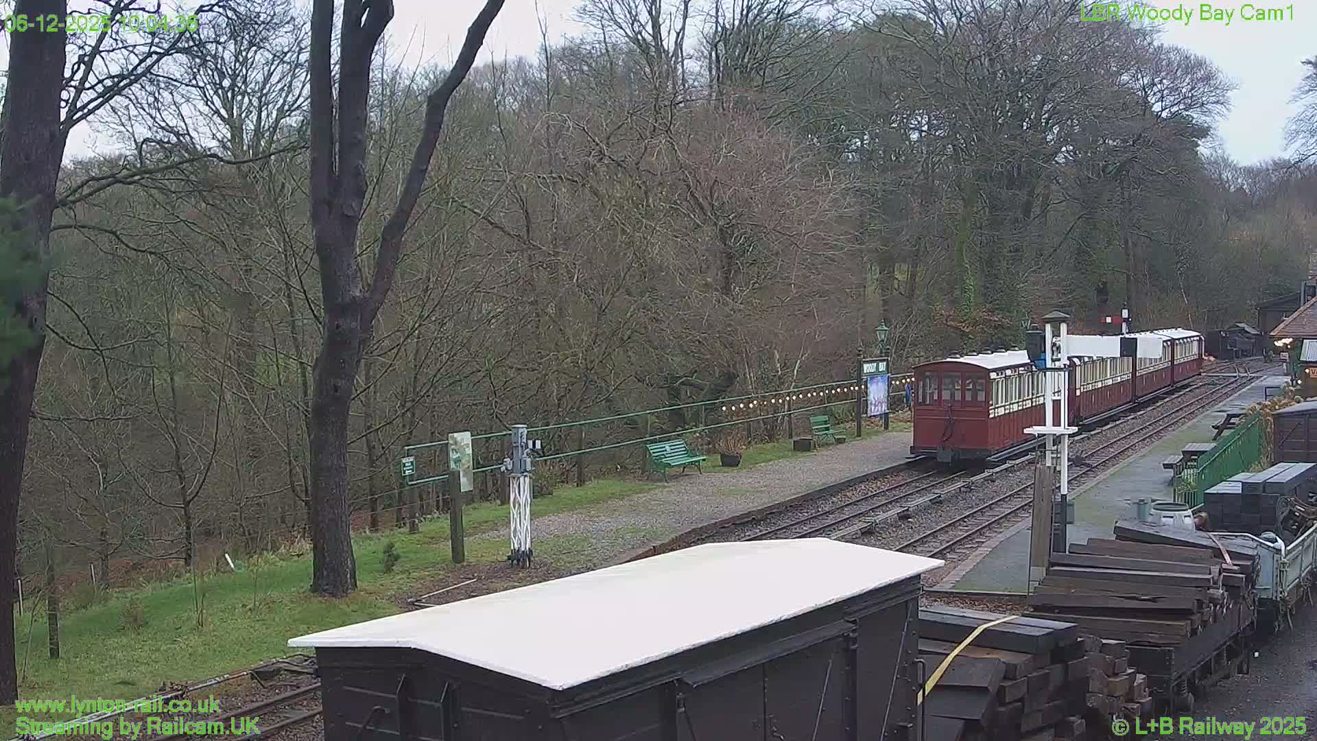 On an overcast day, a red and cream train rests at a scenic railway station with multiple tracks, surrounded by a dense, leafless forest and various railway sidings.