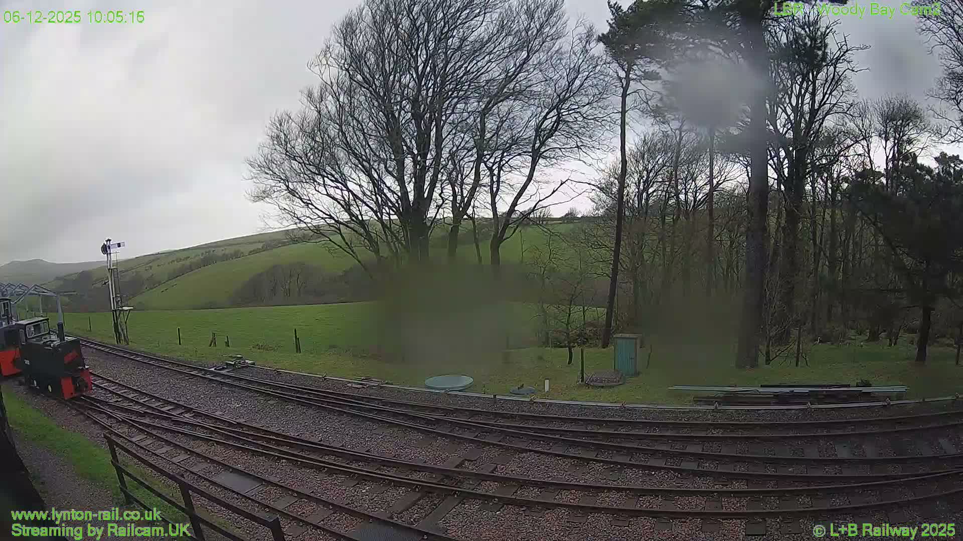 A small train sits on multiple tracks in the foreground, leading towards a signal post, set against a backdrop of green rolling hills and a dense forest of bare trees under a gloomy, overcast sky with visible rain streaks on the camera lens.