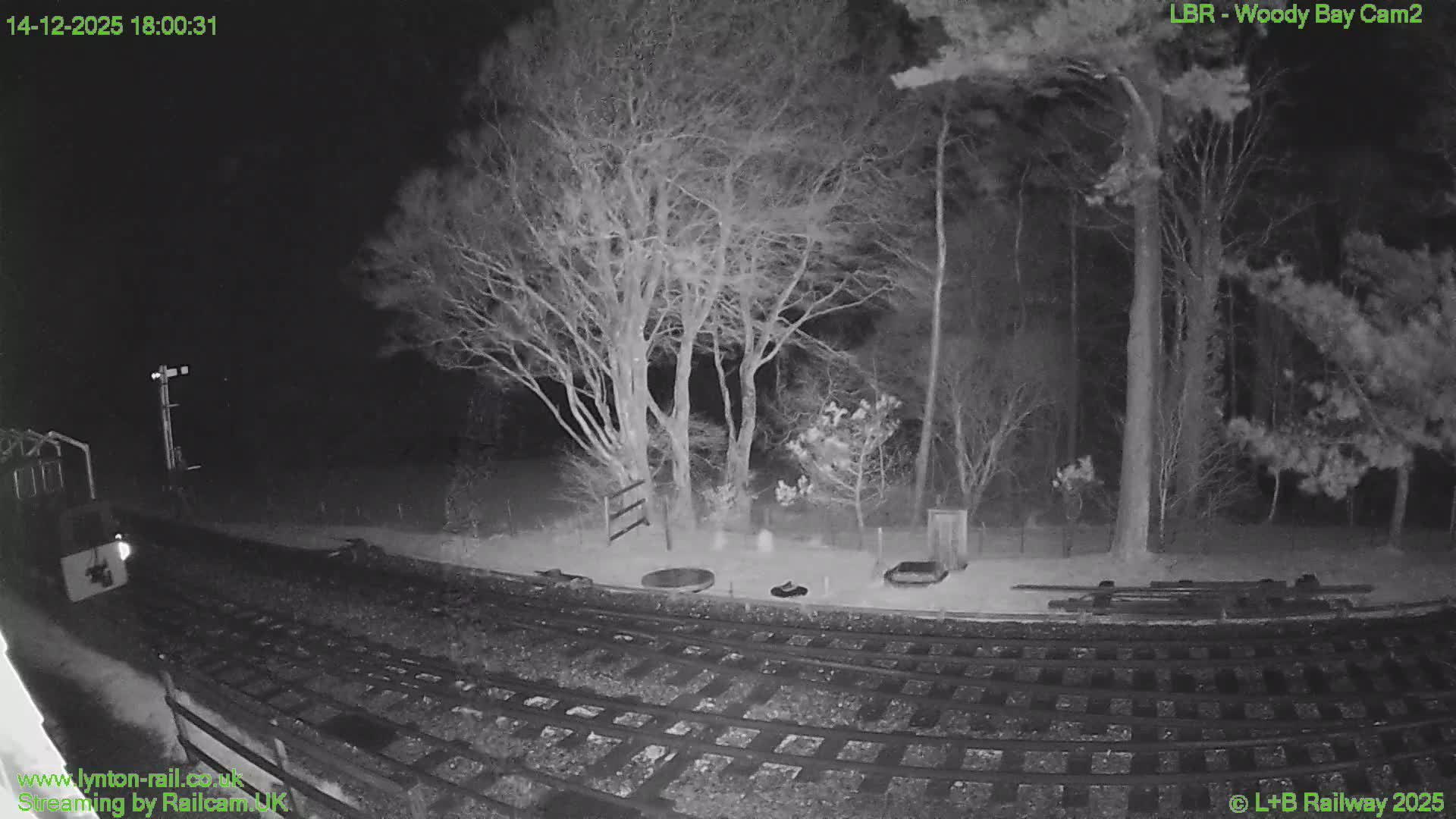 A small train sits on multiple tracks in the foreground, leading towards a signal post, set against a backdrop of green rolling hills and a dense forest of bare trees under a gloomy, overcast sky with visible rain streaks on the camera lens.
