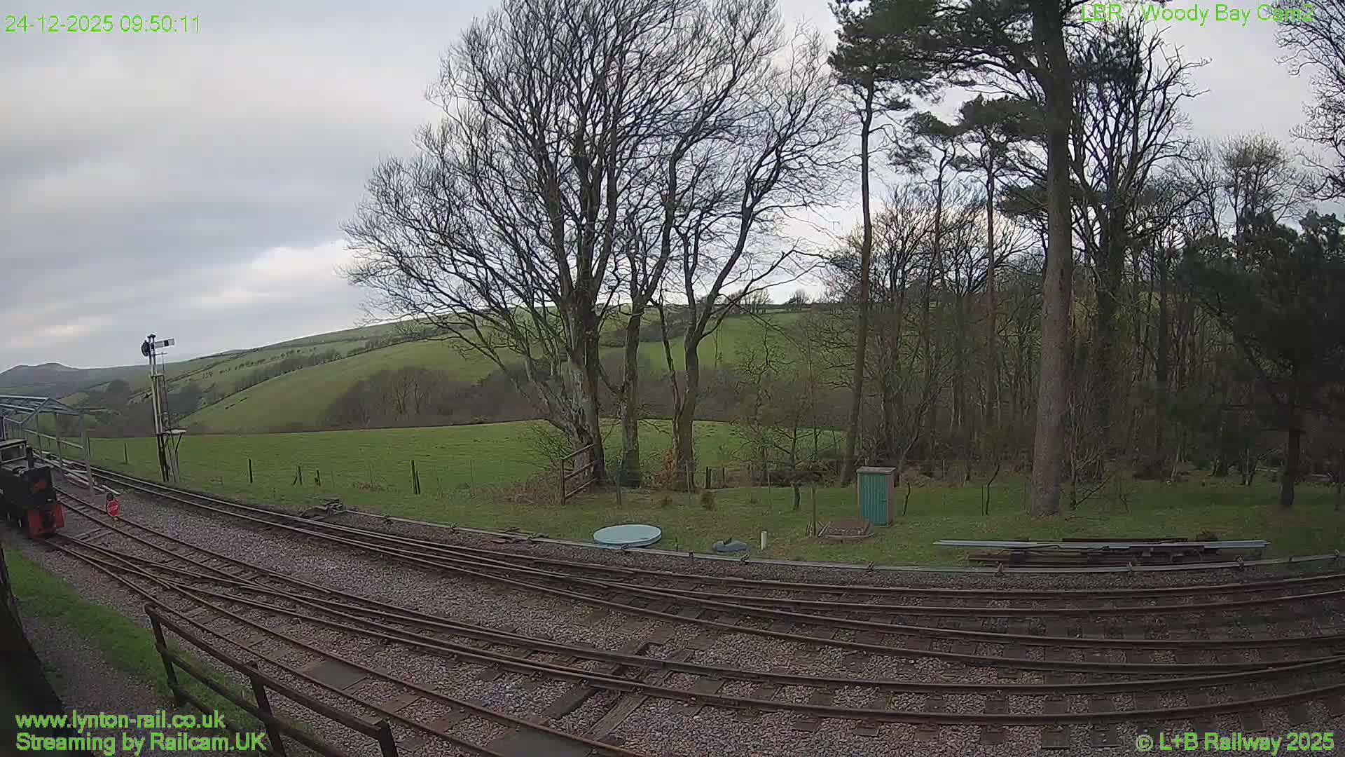 A small train sits on multiple tracks in the foreground, leading towards a signal post, set against a backdrop of green rolling hills and a dense forest of bare trees under a gloomy, overcast sky with visible rain streaks on the camera lens.