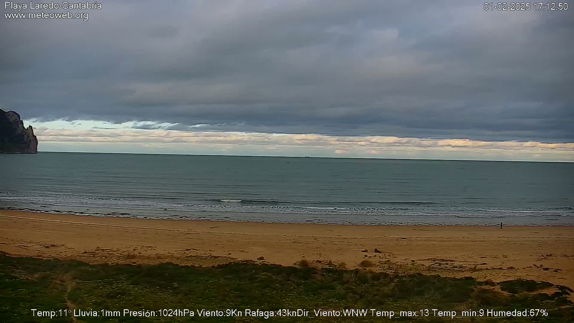A sandy beach with gentle waves laps the shore under a mostly cloudy sky.