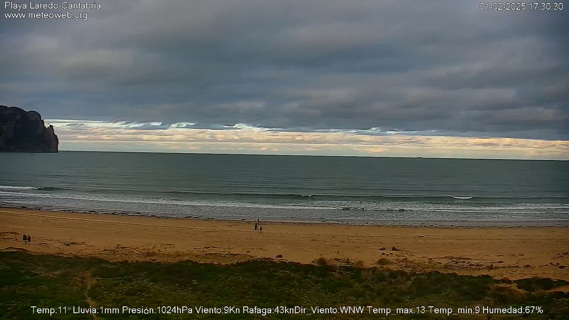 A sandy beach with a few people on it stretches to a calm, gray-green ocean under a mostly cloudy sky.