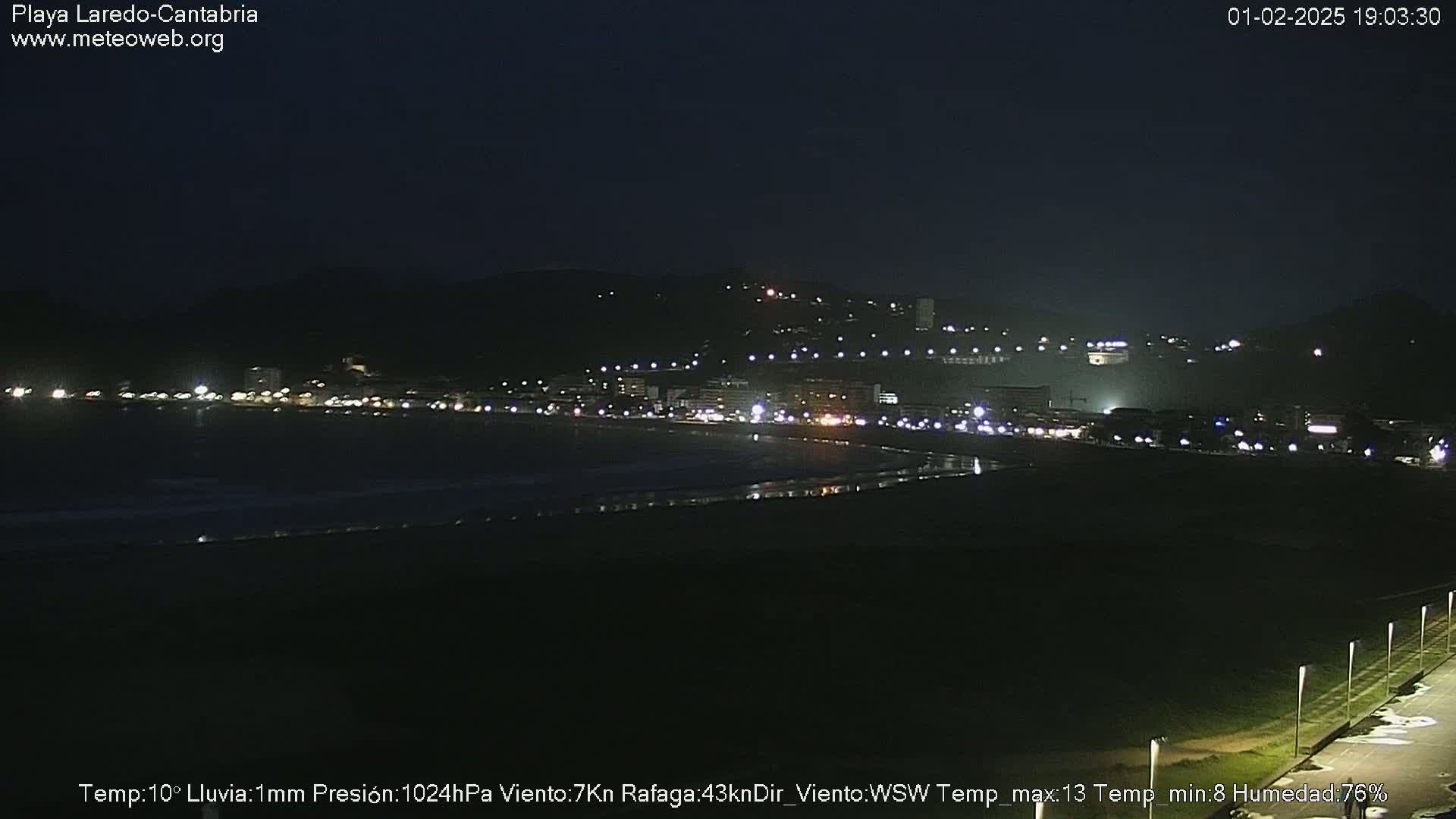 A nighttime view of a dimly lit beach town nestled against a dark hill, under conditions of light rain.