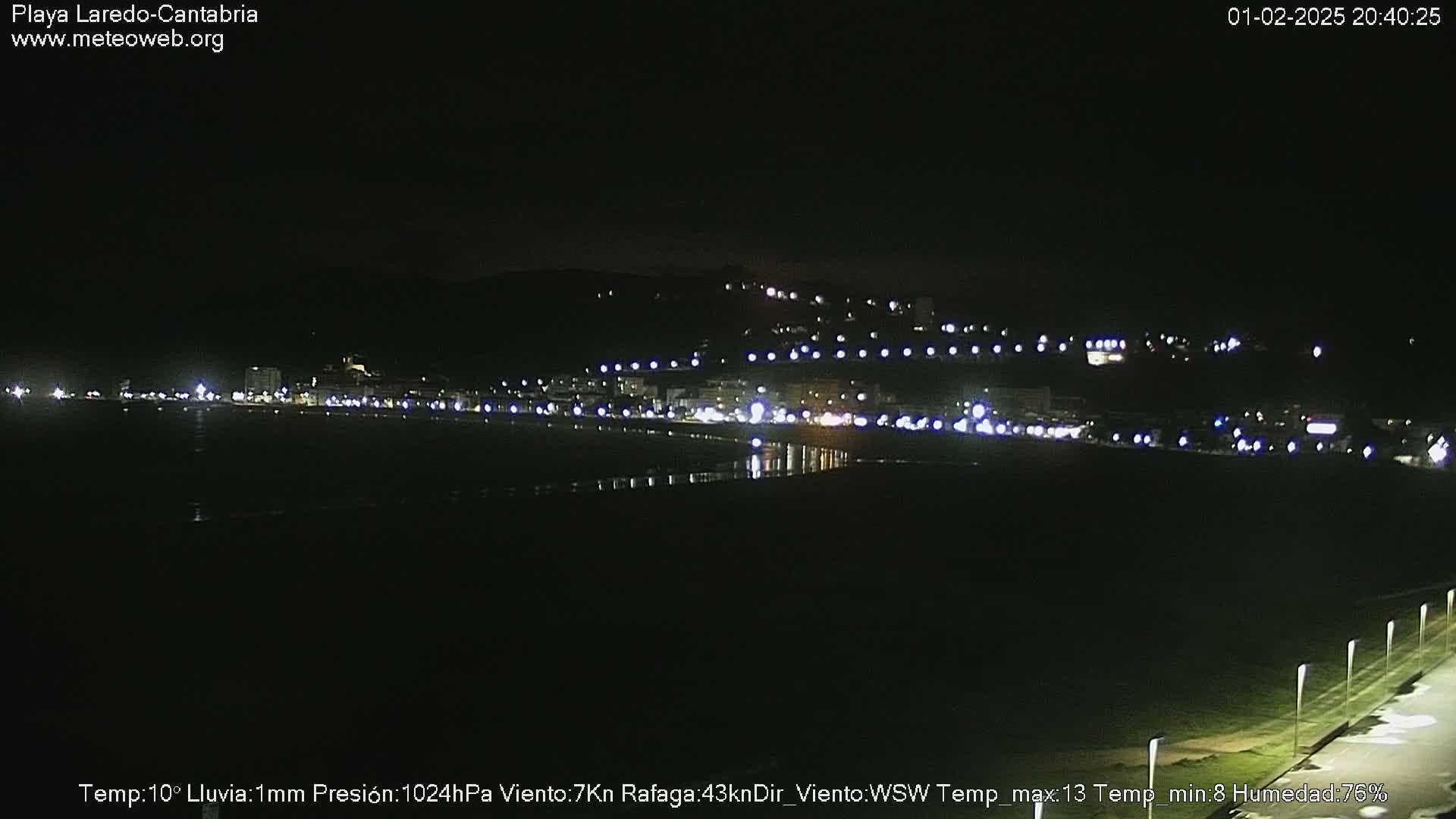 A nighttime view of a coastal town with faint lights illuminating the beach and buildings under a slightly rainy sky.