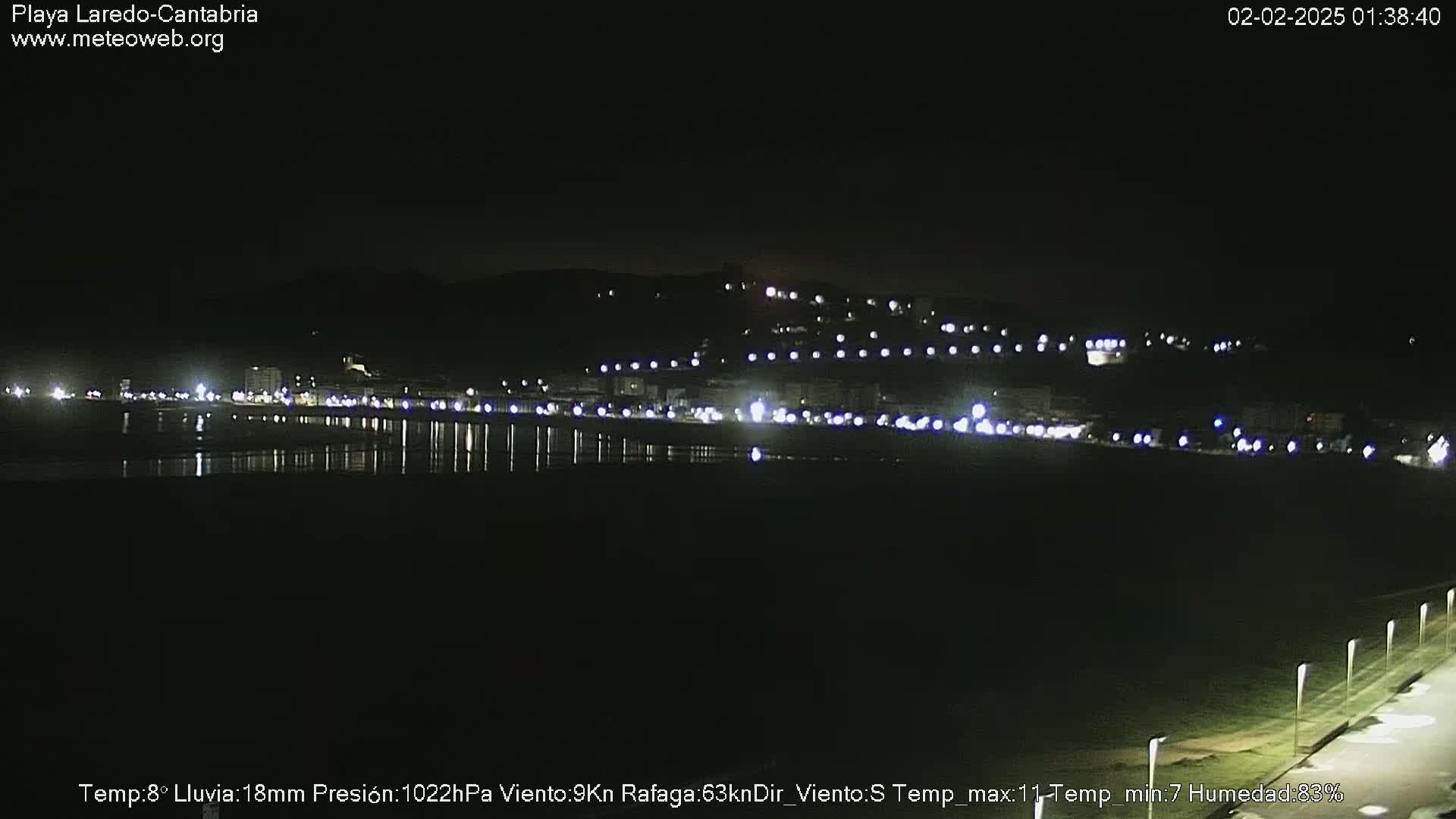 A nighttime view of a calm, dark beach with a town's lights illuminating the shore and a hill in the background.