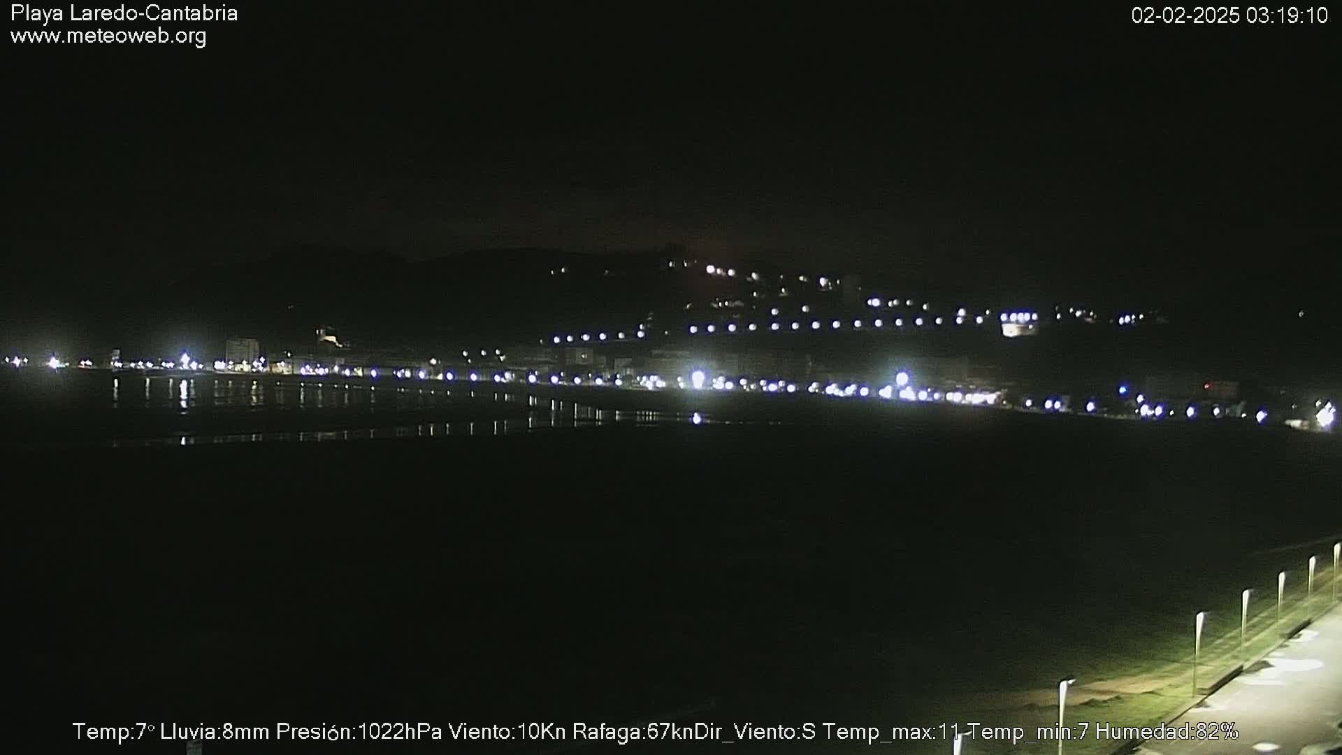 A nighttime view of a coastal town with lights reflecting on a dark body of water under a clear sky.