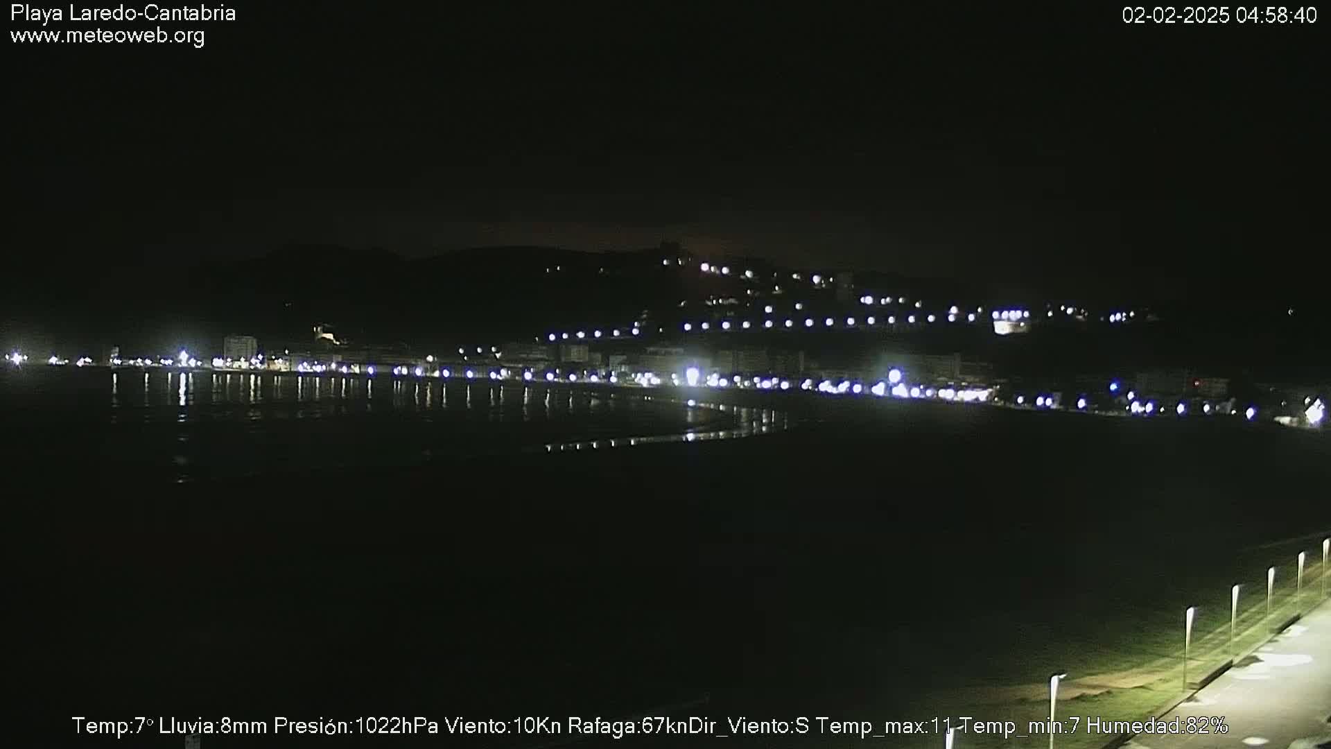 A nighttime view of a coastal town with its lights reflected on the dark water under a clear sky.