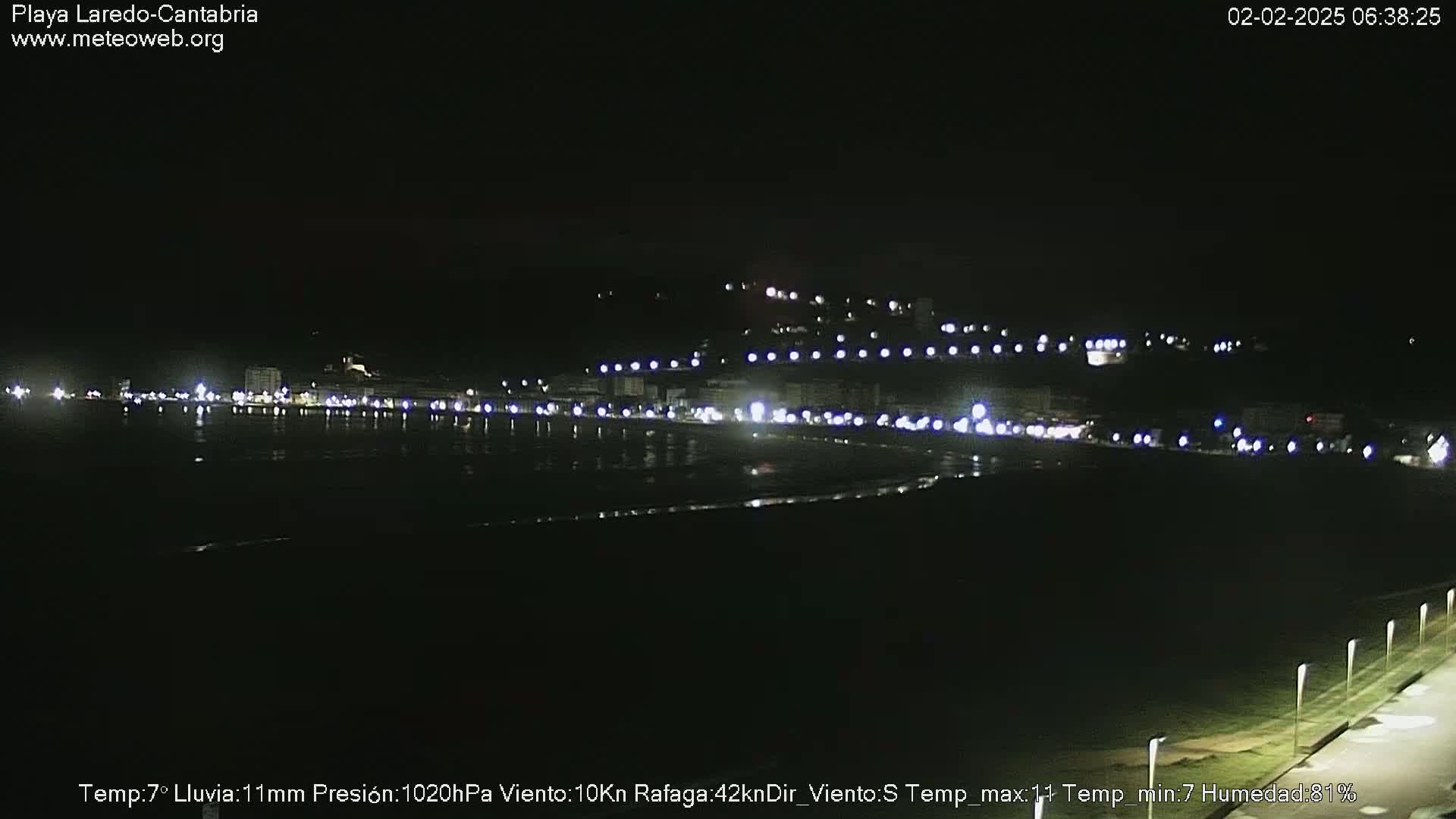 A nighttime view of a dimly lit beach town with streetlights lining the shore, under a dark sky.