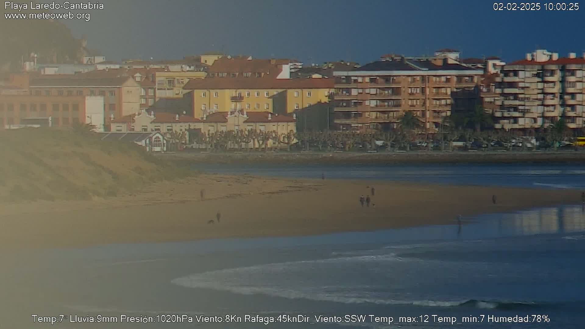 A sandy beach with a few people walking on it, overlooks a calm ocean with small waves, and a town of multi-story buildings in the background under a clear sky.