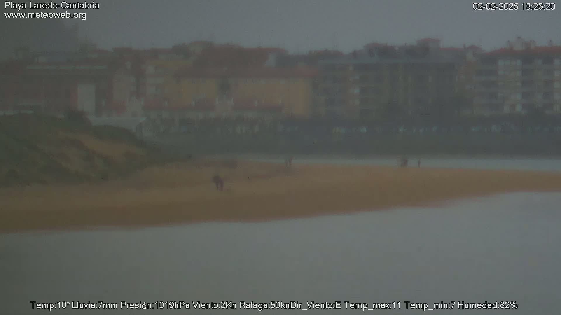 A hazy, rainy view of a beach with a few people visible in the distance and apartment buildings in the background.