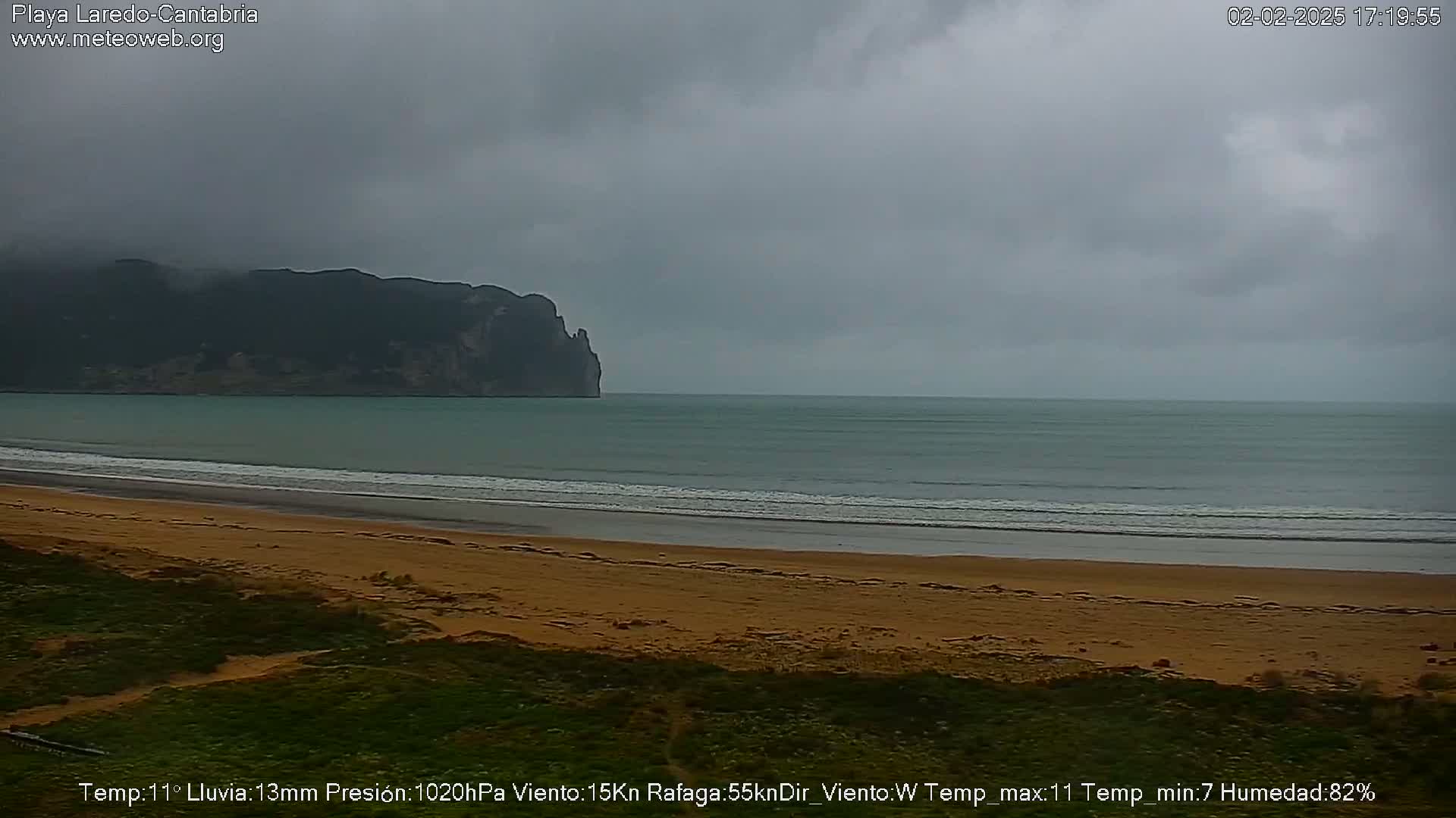 A sandy beach stretches to a calm ocean under an overcast sky, with a dark mountain visible in the distance.