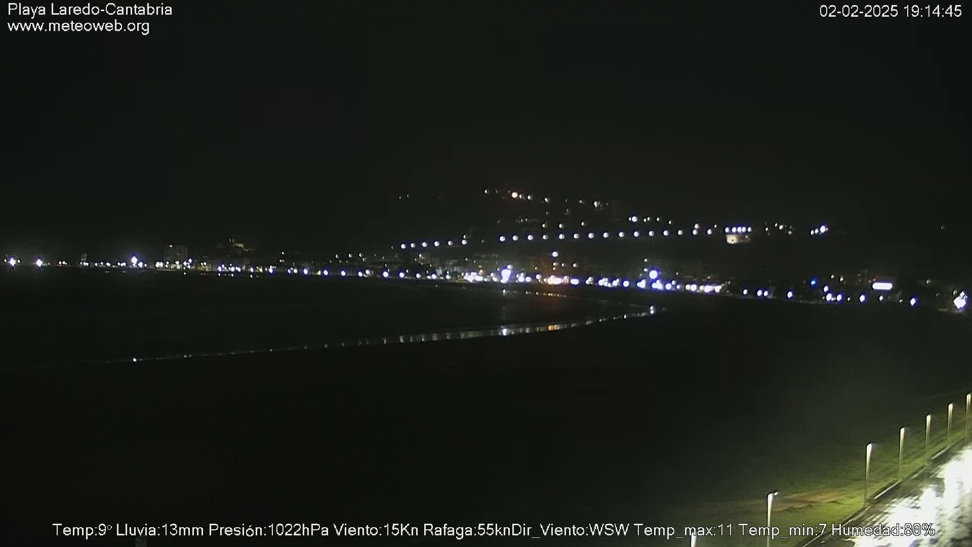 A nighttime view of a dark, sparsely lit beach with a town's lights visible in the distance under a clear, dark sky.