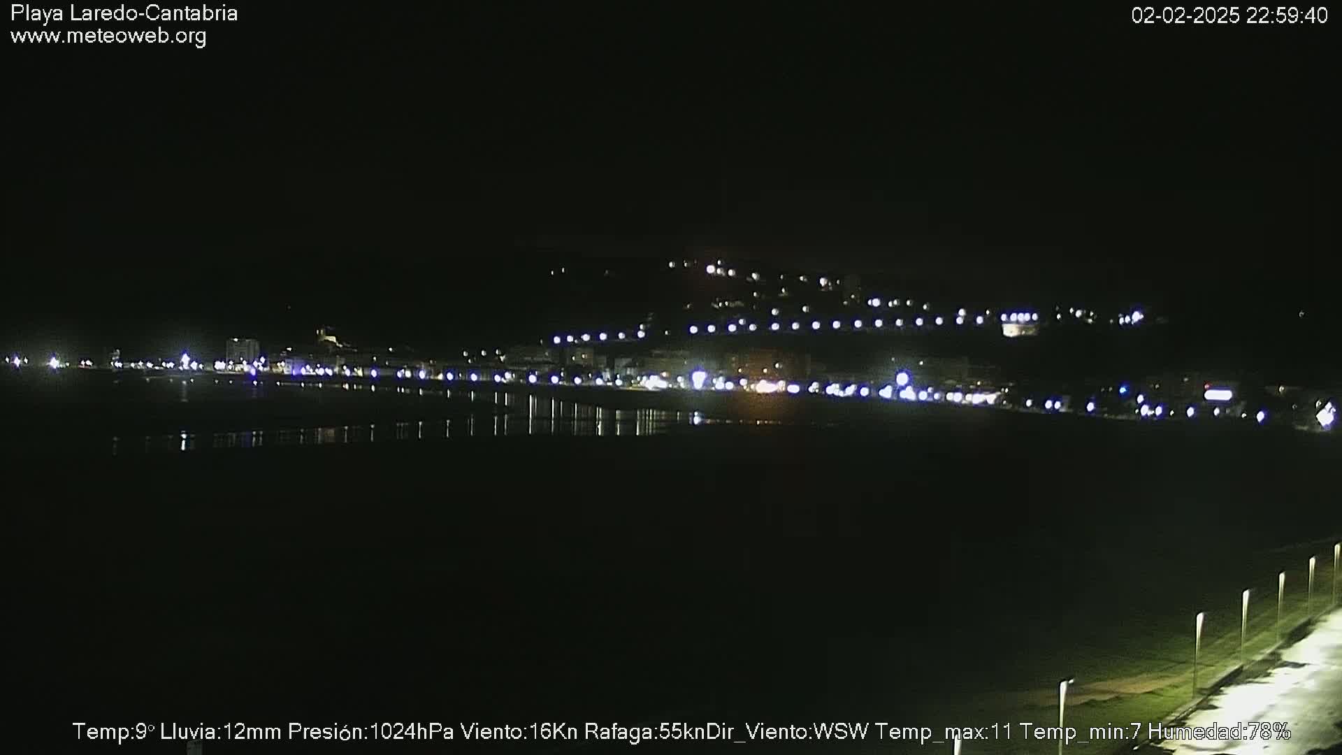 A nighttime coastal town is illuminated with lights, reflecting in dark water under a dark, rainy sky.