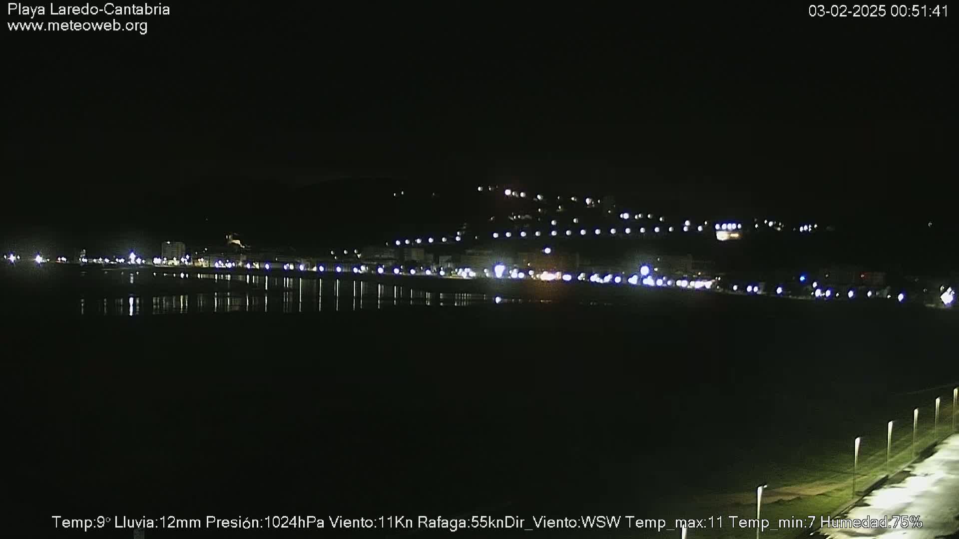 A nighttime view of a coastal town with its lights reflected in still water under a dark, rainy sky.