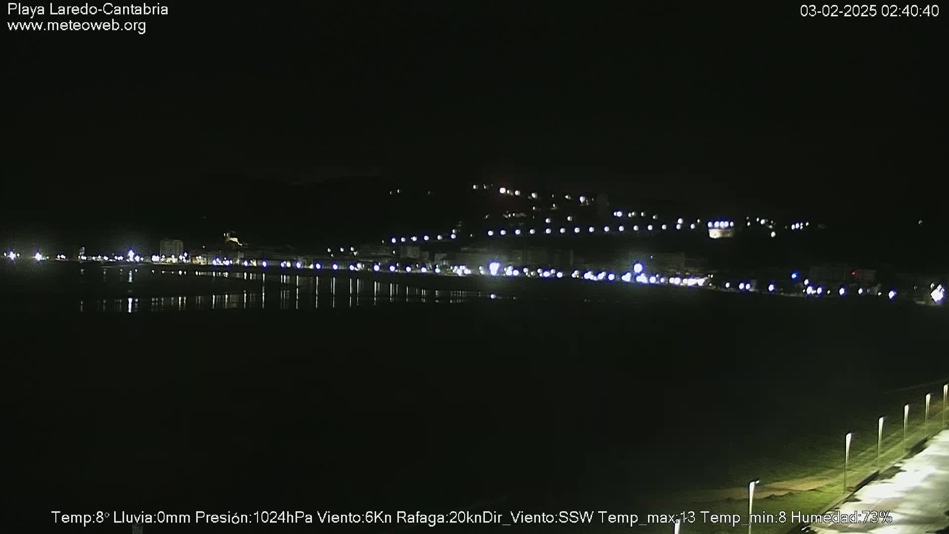 A nighttime view of a coastal town illuminated by lights, reflecting on dark water under a clear sky.