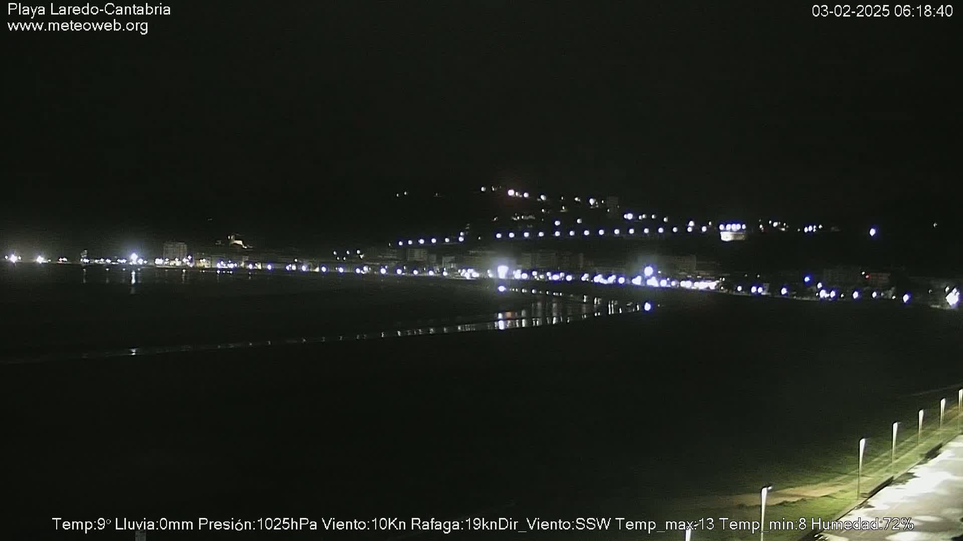 A nighttime view of a coastal town with faint lights and a dark, possibly wet, beach.