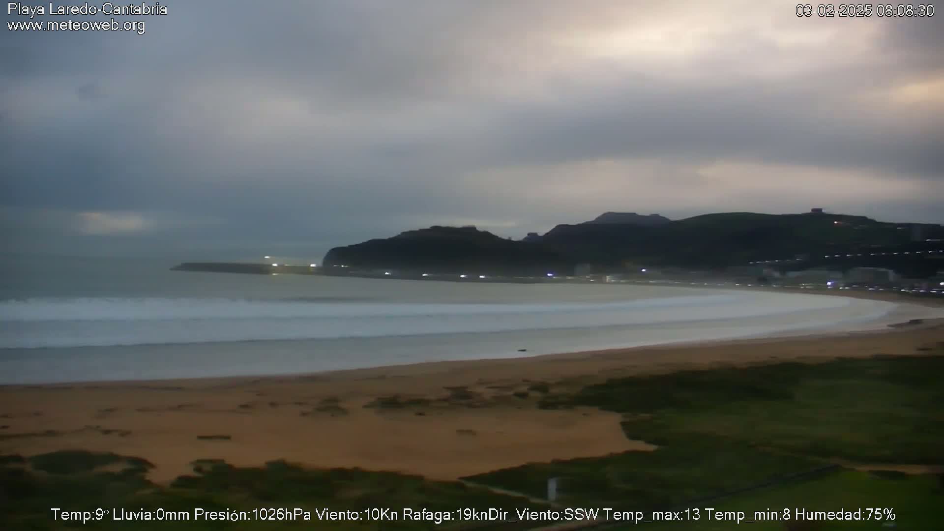A wide sandy beach with gentle waves stretches towards a dark, hilly coastline under an overcast sky.