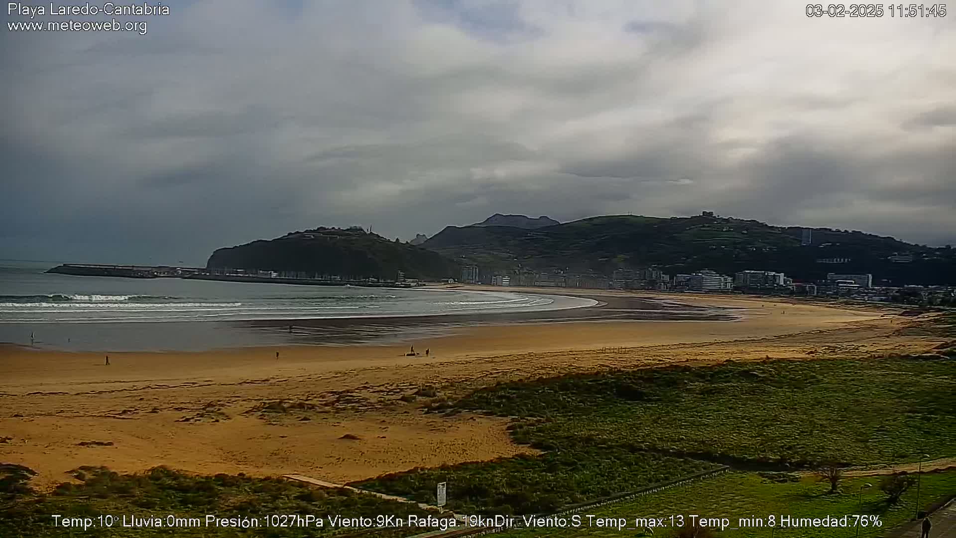 A long sandy beach with gentle waves, a town in the background, and overcast skies stretches out before a low hill.