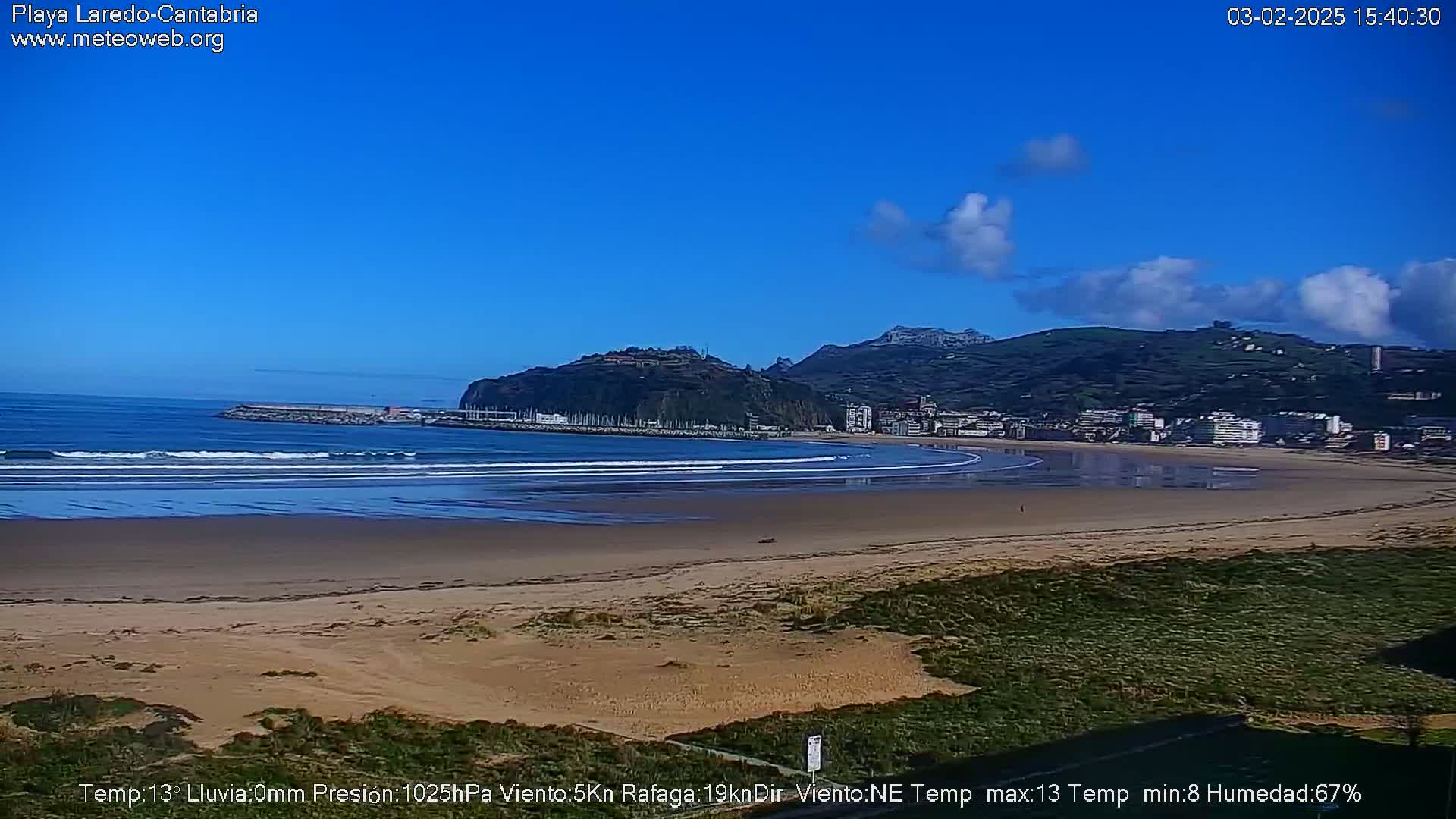 A sandy beach with gentle waves stretches out to a town nestled against a verdant hillside under a mostly clear blue sky.