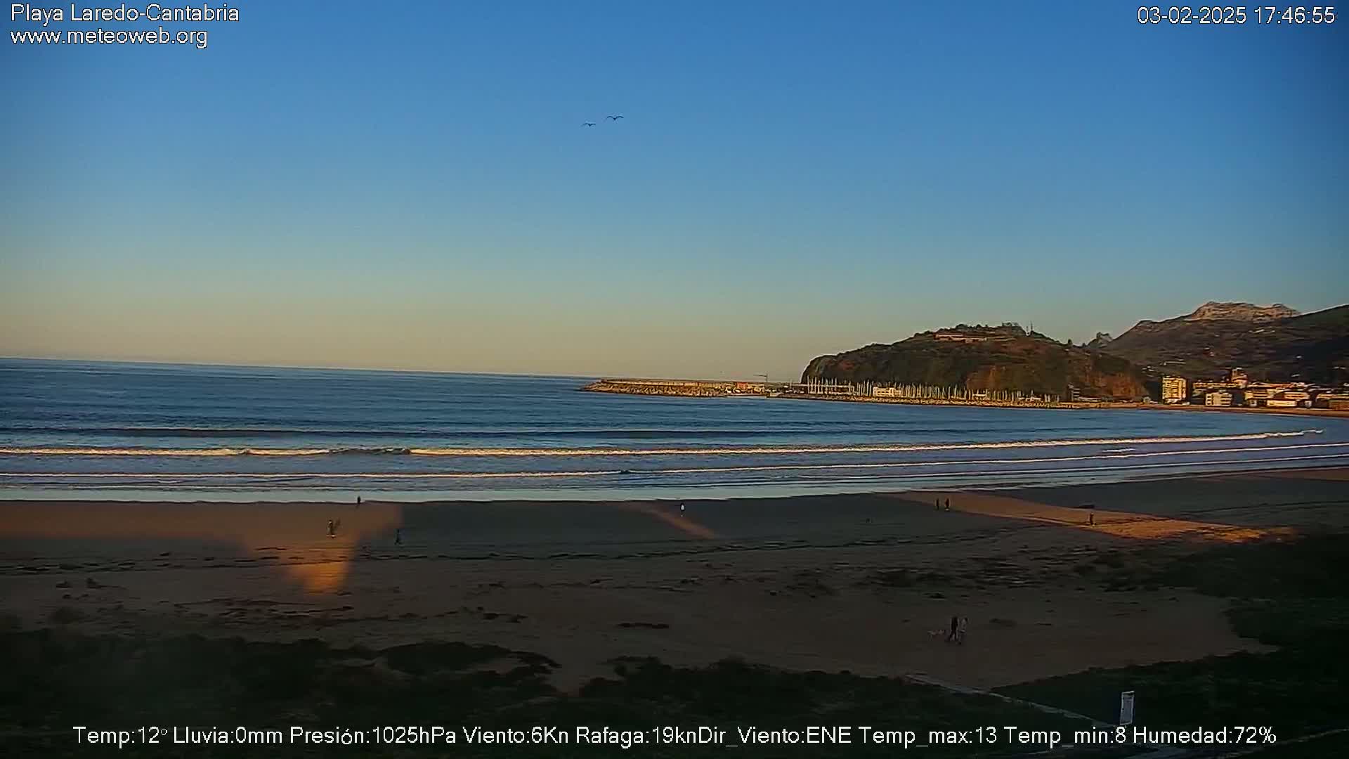 A sandy beach with gentle waves under a clear, twilight sky, backed by a calm harbor and distant hills.