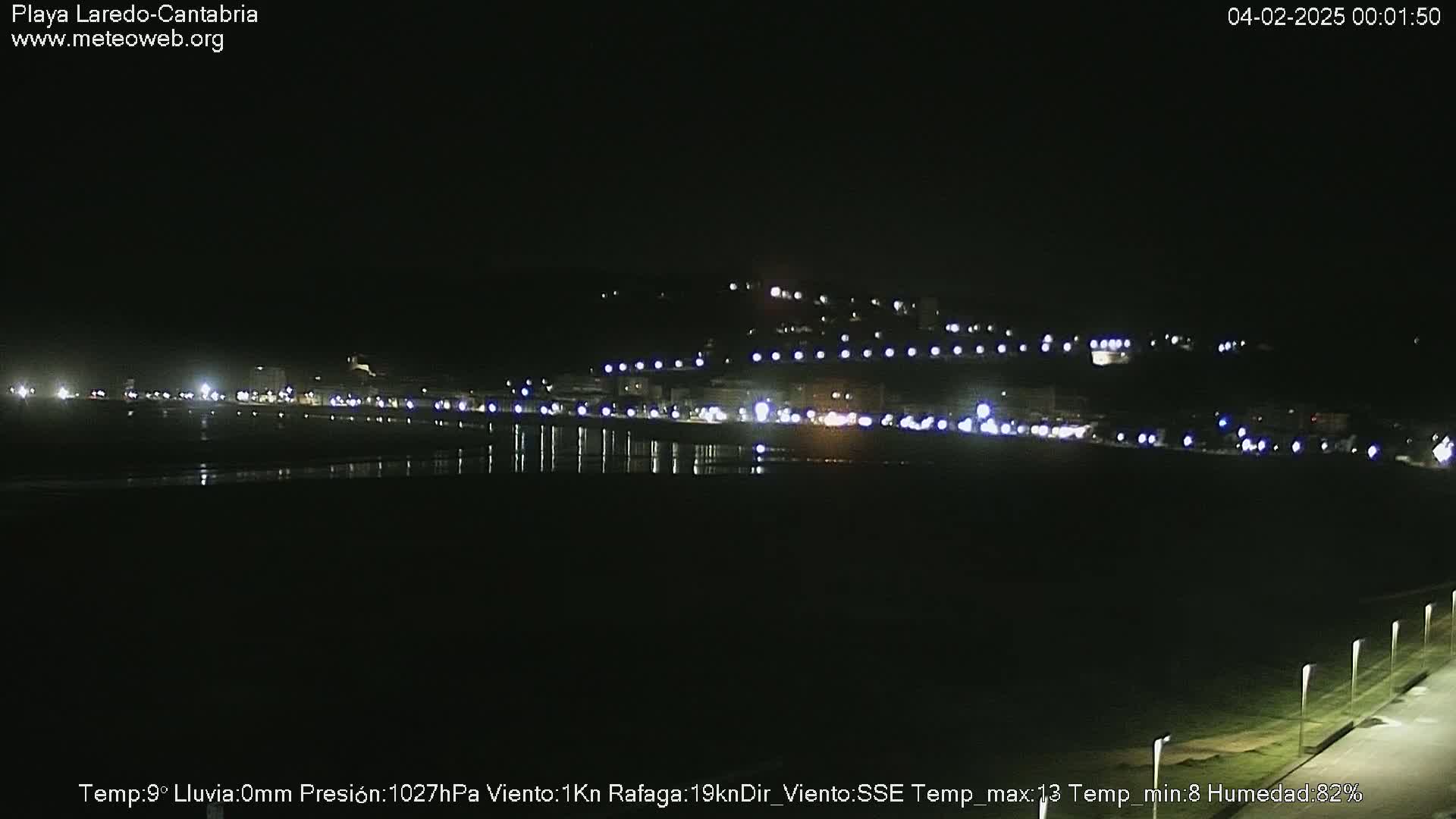 A nighttime view of a coastal town with lights reflecting on a calm body of water under a clear, dark sky.