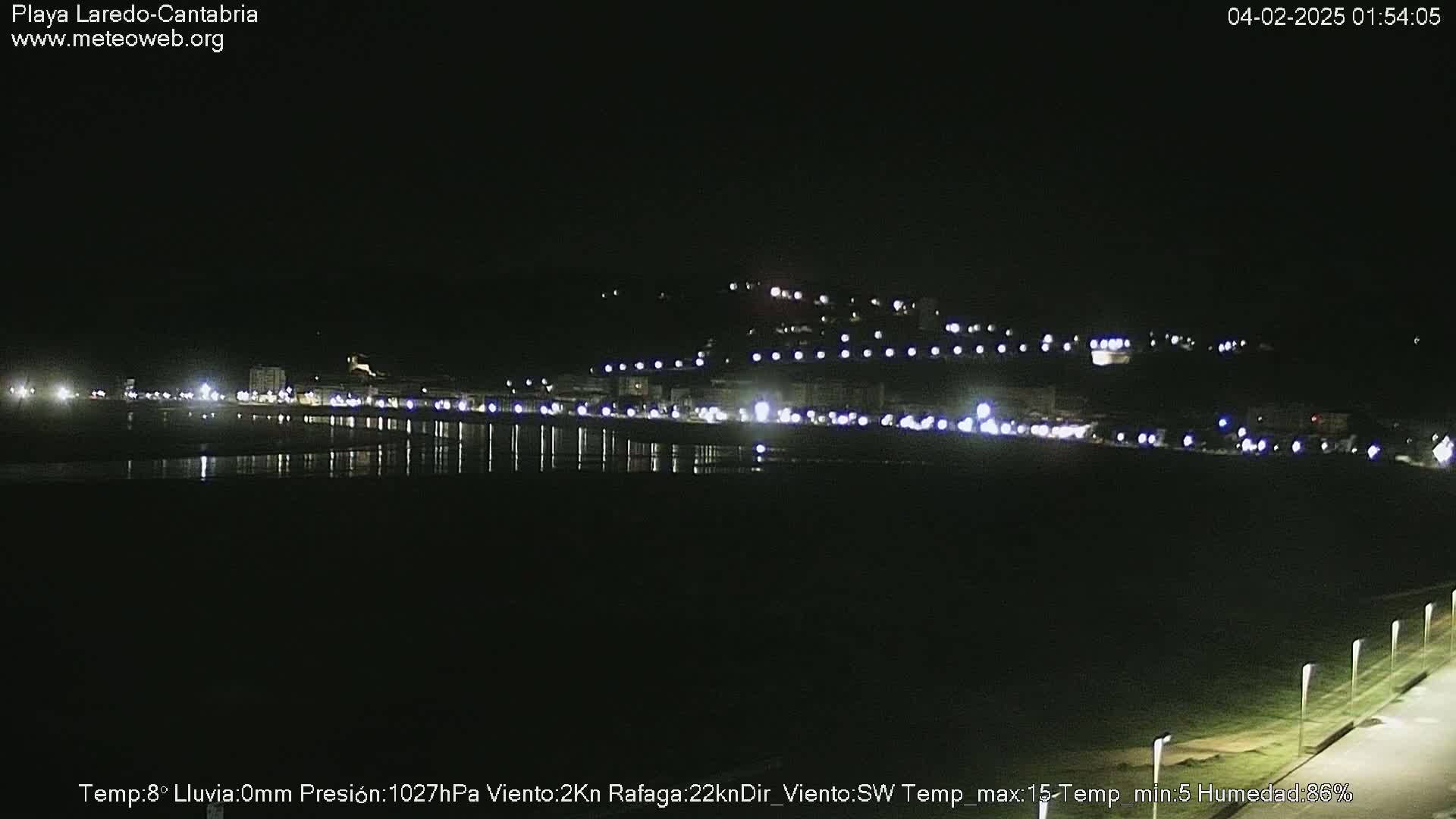 A nighttime view of a coastal town with lights reflecting on a dark body of water under a clear, dark sky.