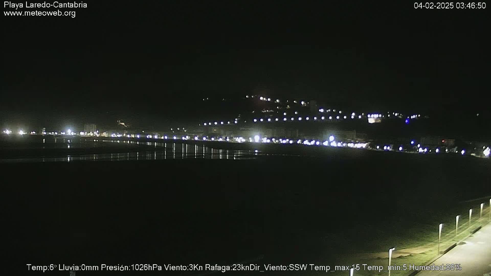 A nighttime view of a dimly lit beach and town with a dark, calm ocean and lights visible on the shore and in the distance.