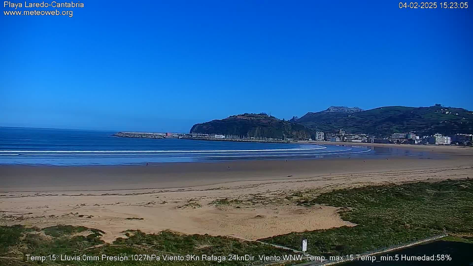 A long sandy beach stretches out to a calm ocean under a clear blue sky, with a town and hills visible in the distance.