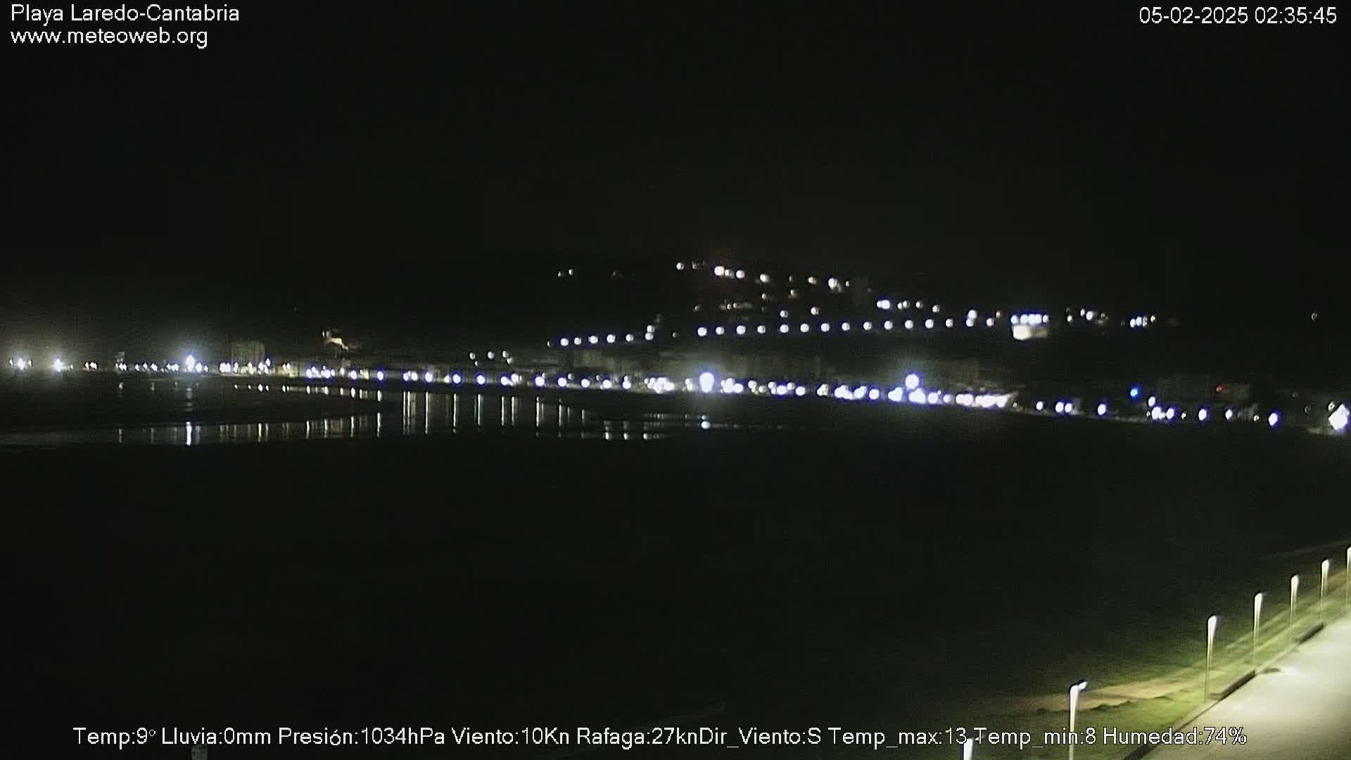 A nighttime view of a dark beach and a town illuminated by lights, under clear skies.