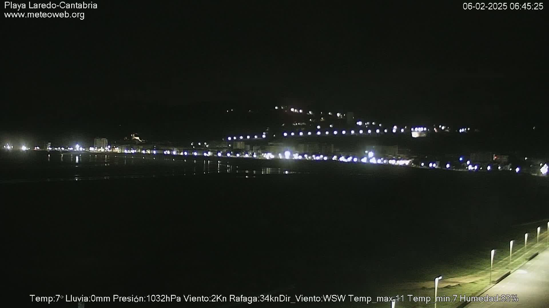 A nighttime view of a coastal town with lights reflecting on a dark body of water, under a clear sky.