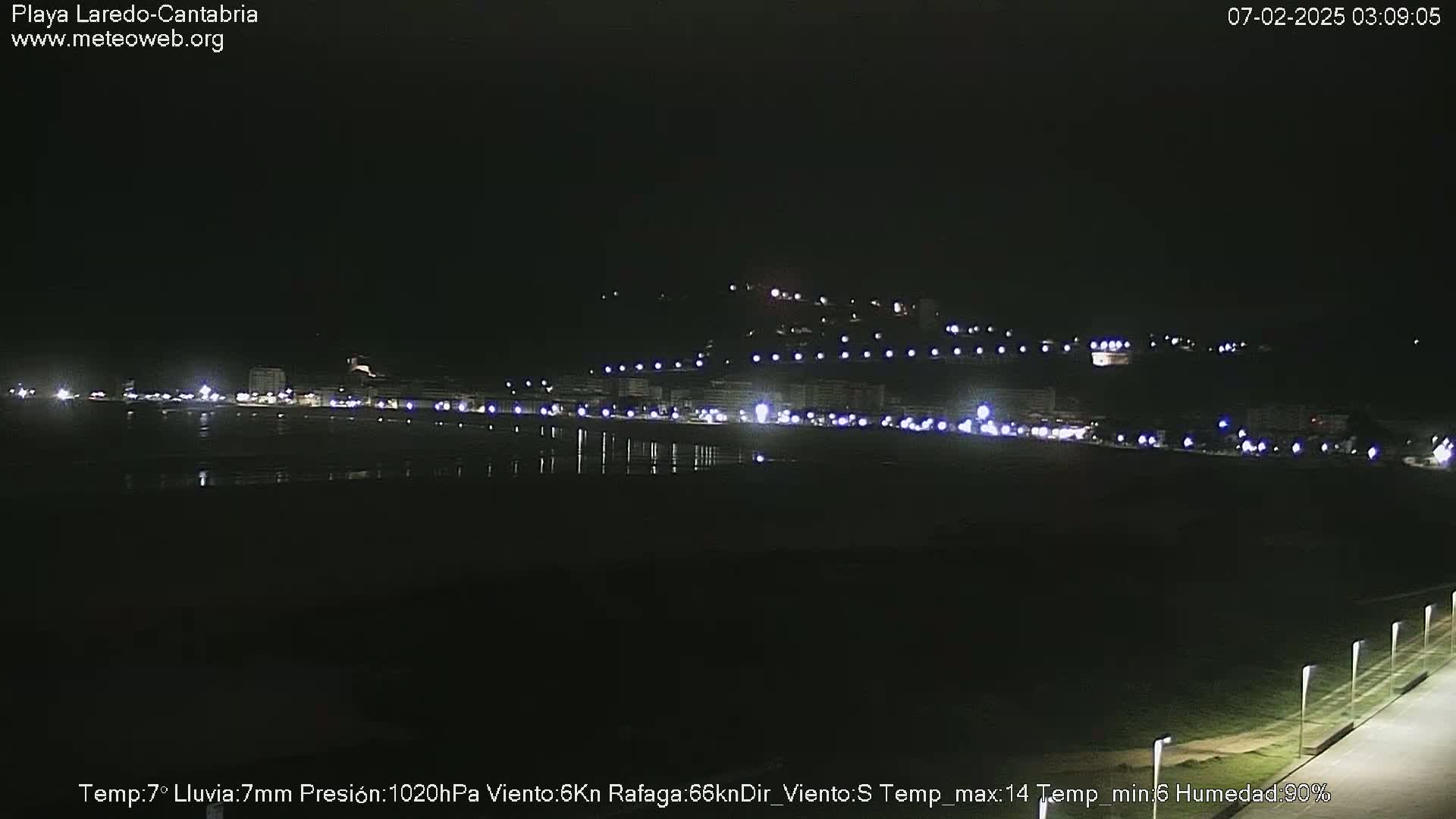 A nighttime view of a dimly lit beach and buildings, with faint lights visible along the shoreline under a dark sky.