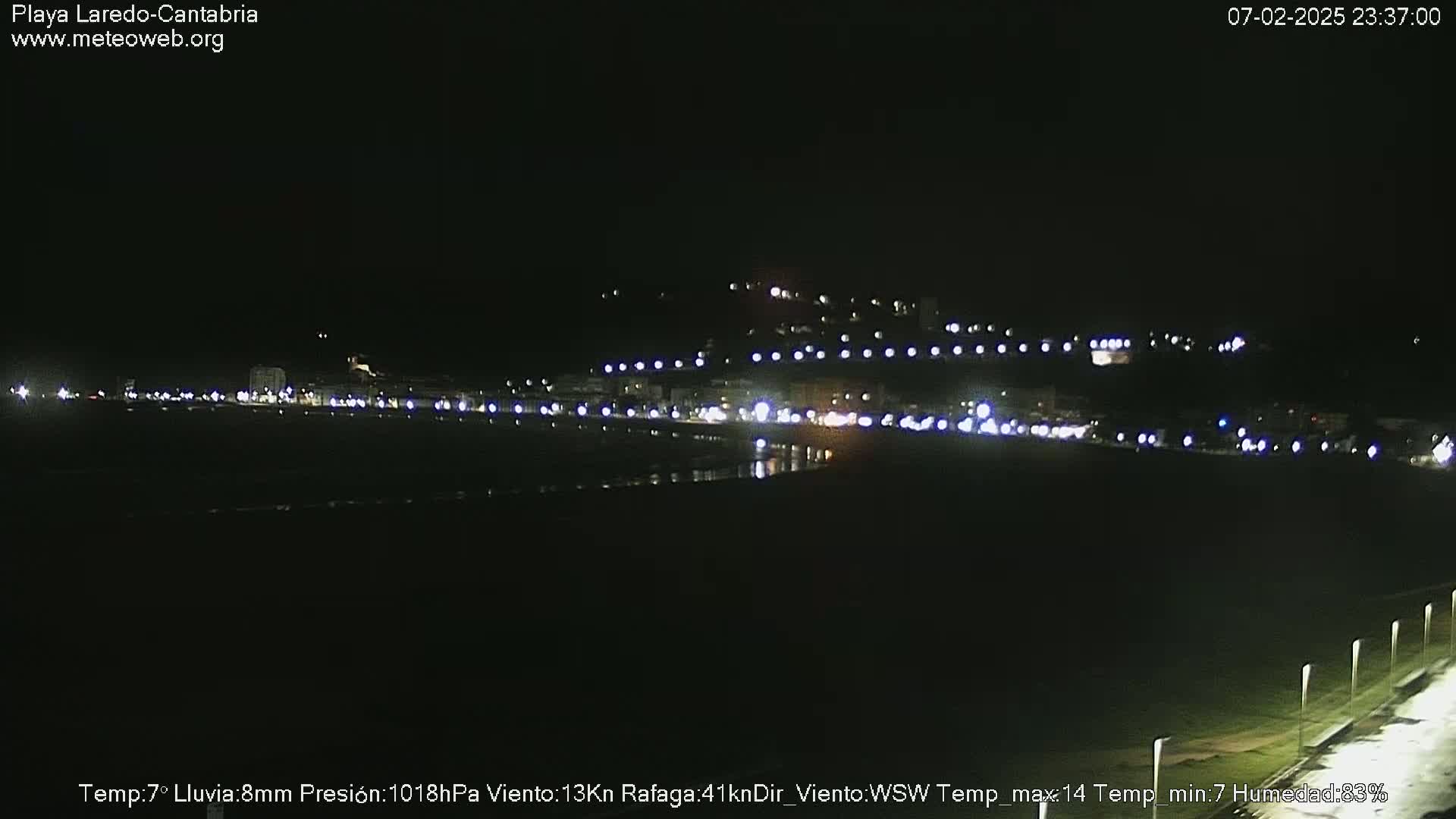 A nighttime view of a coastal town with its lights illuminating the beach and surrounding hills under rainy conditions.