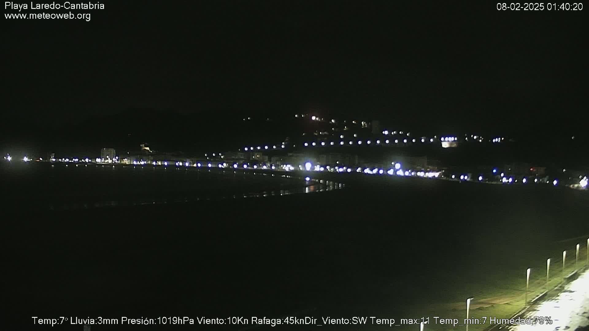 A nighttime view of a dimly lit beach town, with faint lights reflecting on the dark water under a clear sky.
