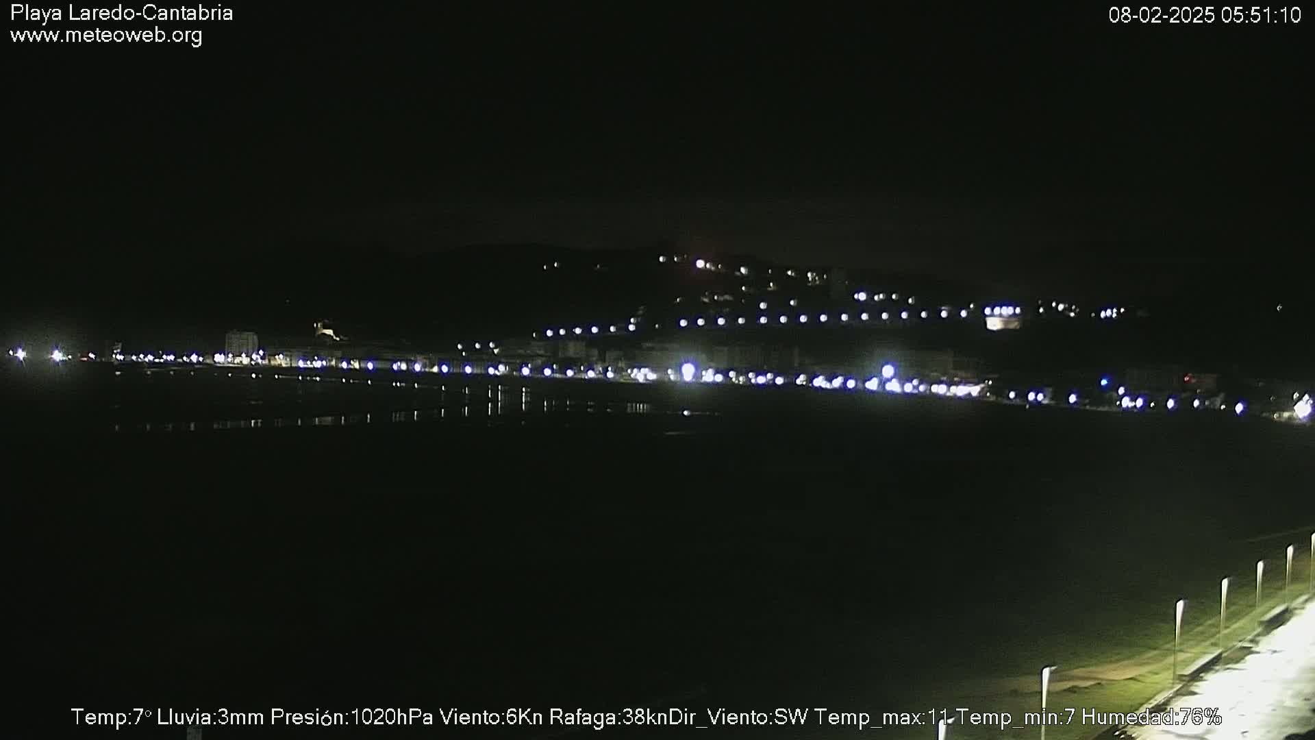 A nighttime view of a dimly lit coastal town, with a dark beach and a dark body of water in the foreground, under a clear, dark sky.